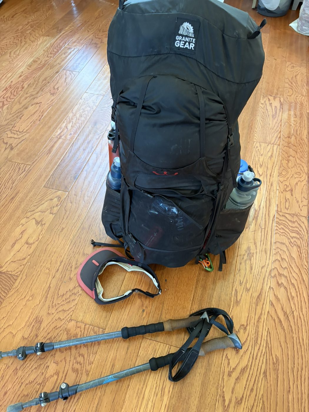 A packed black Granite Gear backpack standing upright on a wood floor with trekking poles and sunglasses beside the pack