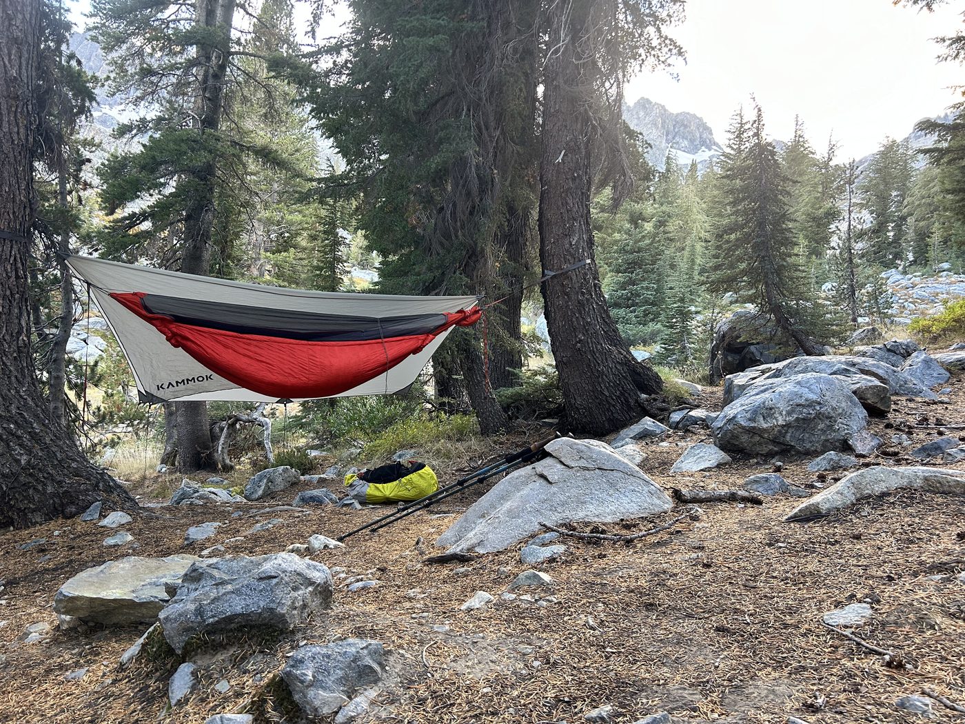 A red hammock with a gray rainfly marked Kammok strung between two pines above a rocky campsite, with mountains behind