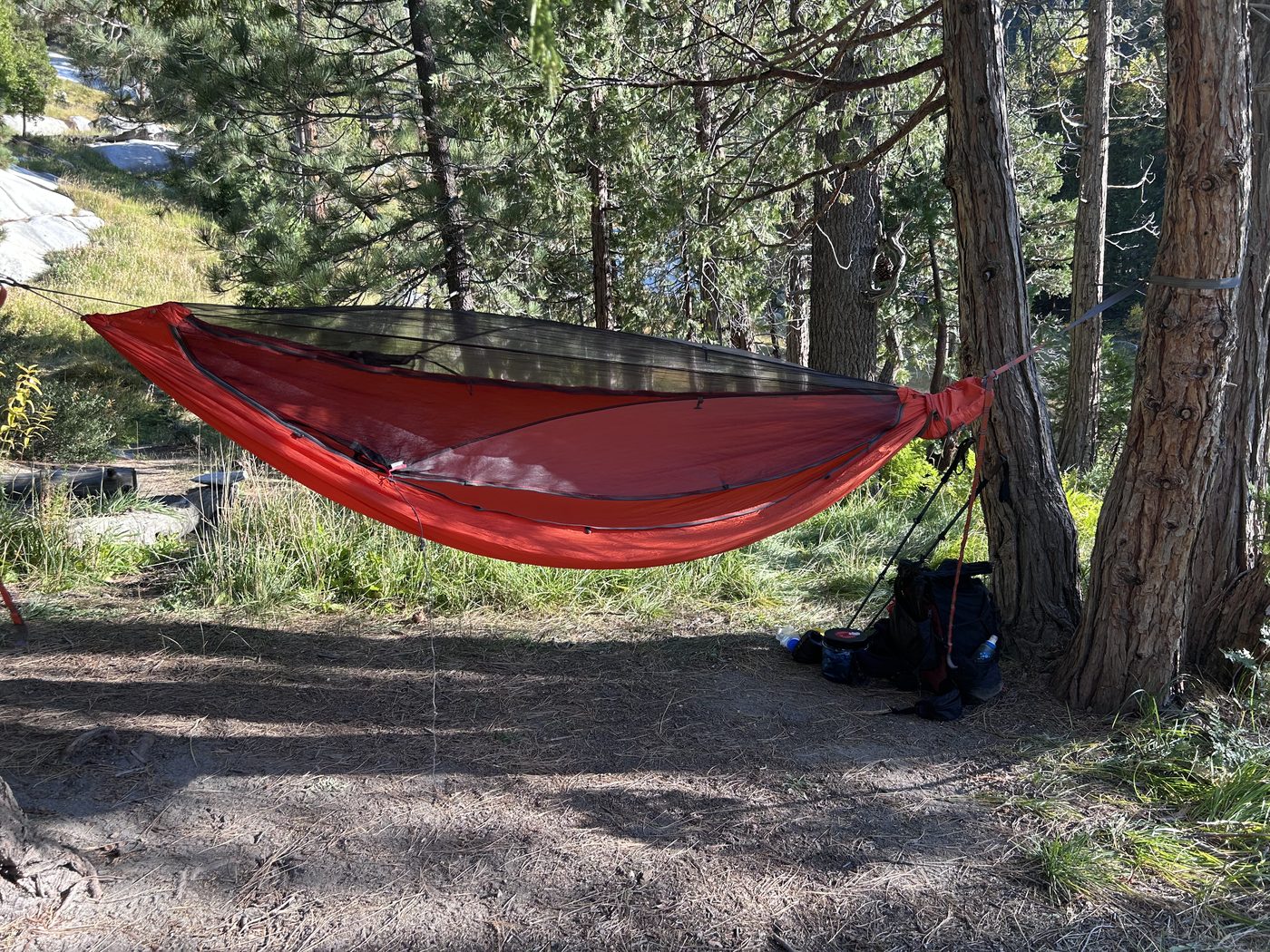 A red hammock with a mesh bug net hung by straps between two pine trunks in a shaded forest camp