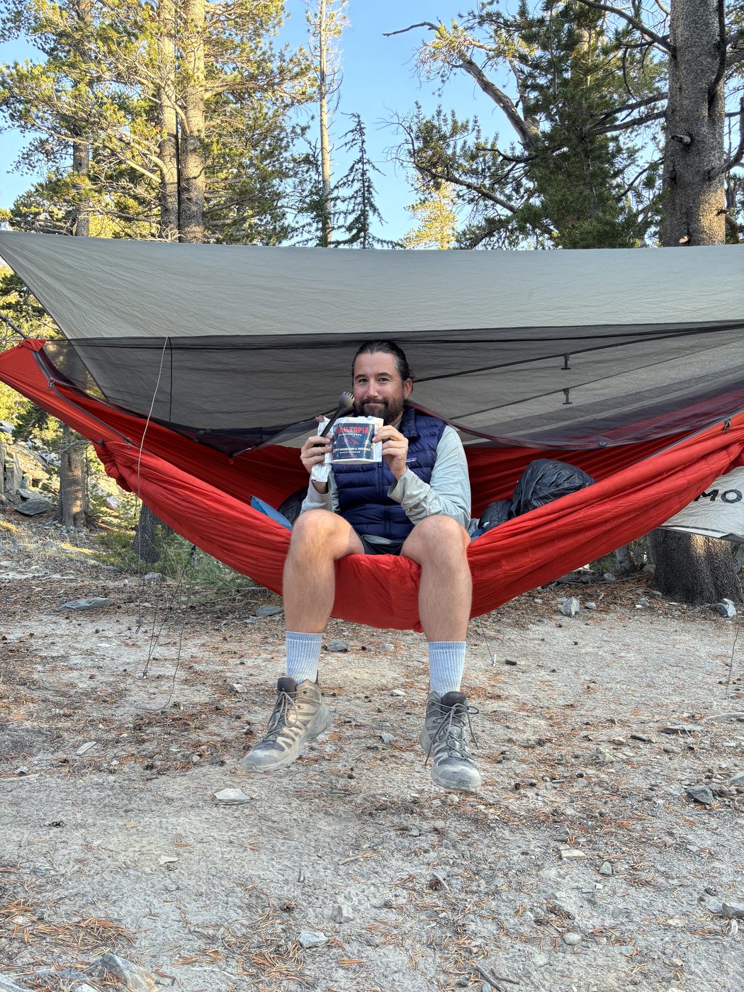 A man in a navy down vest sitting in a red hammock under a gray rainfly at a forest camp