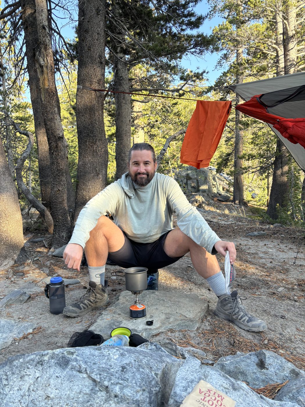 A man crouched on a granite slab beside a small canister stove and a pot, with a hammock and rainfly strung in the trees behind