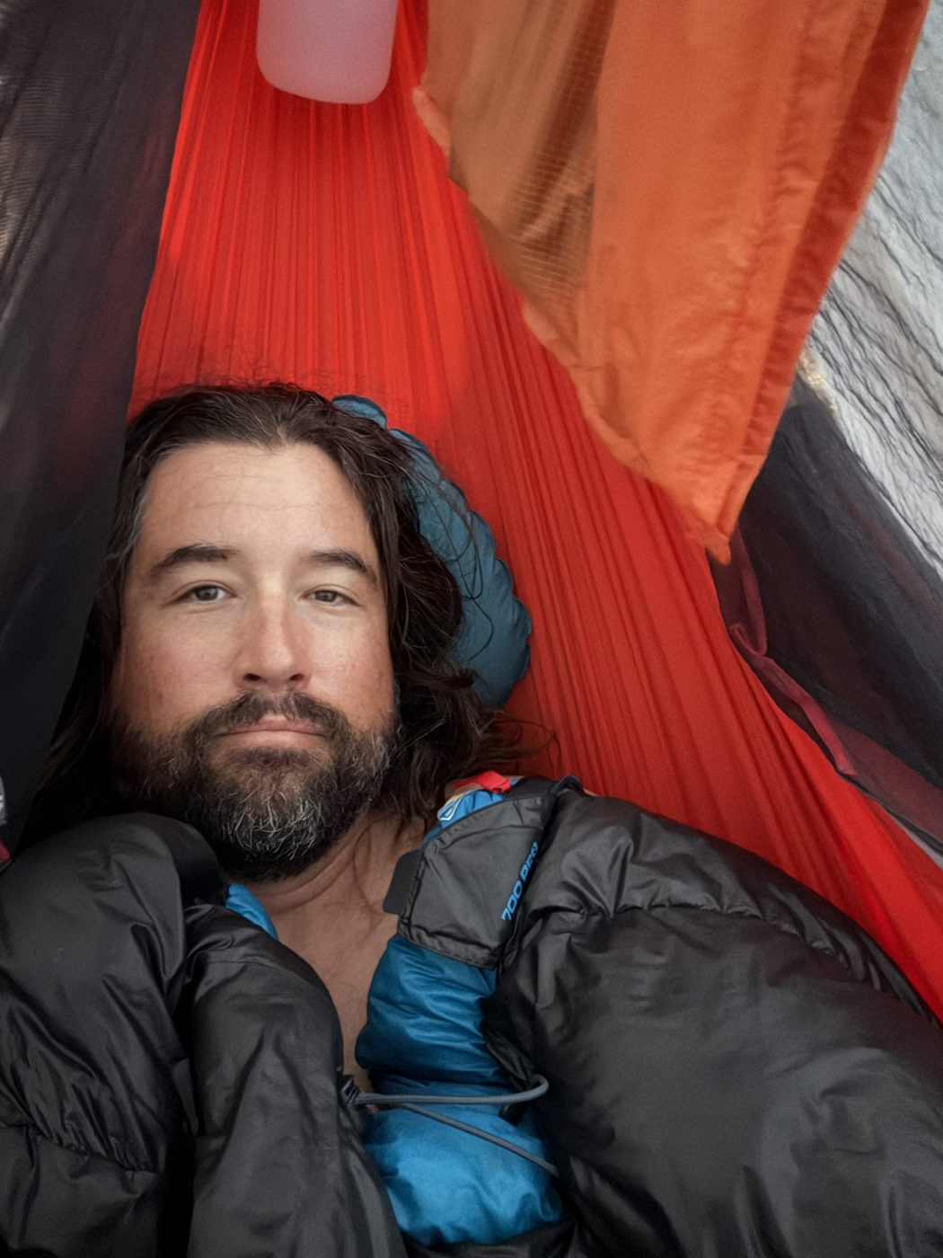 A bearded man lying inside a red hammock with a dark sleeping bag draped over him as a quilt