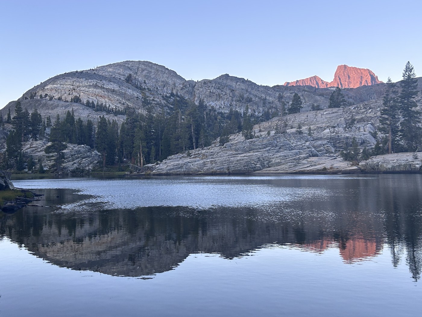 A mirror-flat lake at dawn reflecting low granite ridges and dark pines, with a distant high peak lit orange by the first sun while the foreground stays in shadow