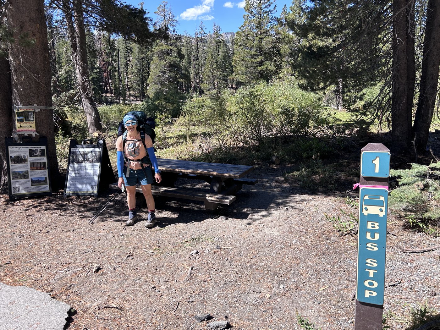 A backpacker standing beside a blue shuttle bus stop post at a forested trailhead, pack still on and trekking poles in hand