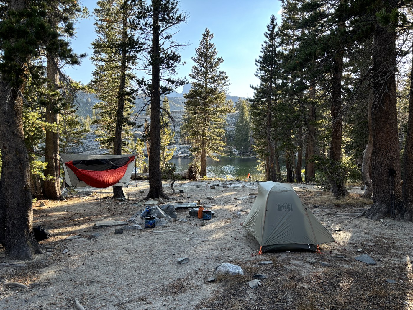 A camp among pines beside a lake, with a hammock strung under a gray tarp and a small green tent pitched on bare ground nearby