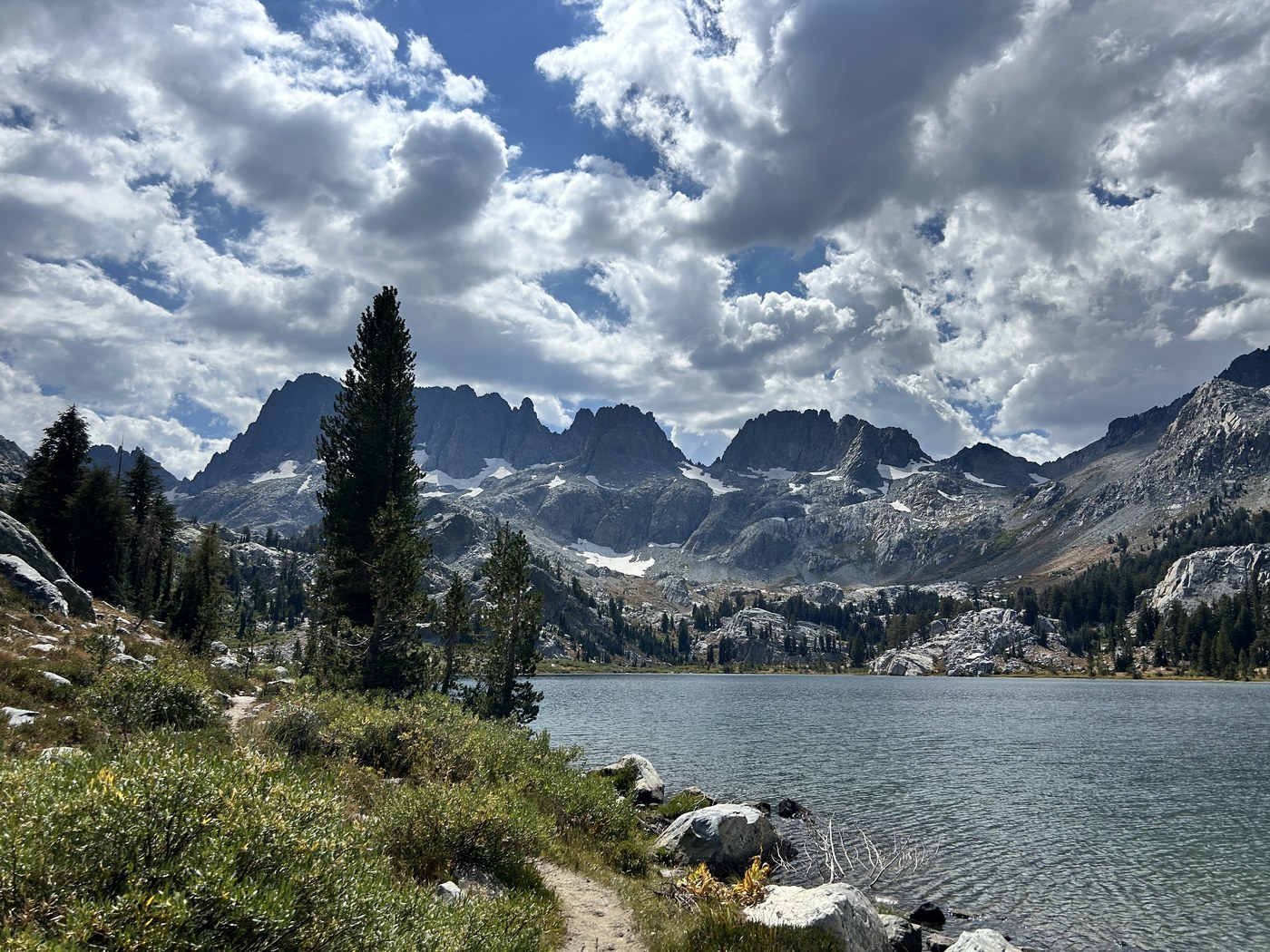 A clear alpine lake in the foreground with a long serrated wall of dark granite spires standing behind, patches of snow lying in the gullies