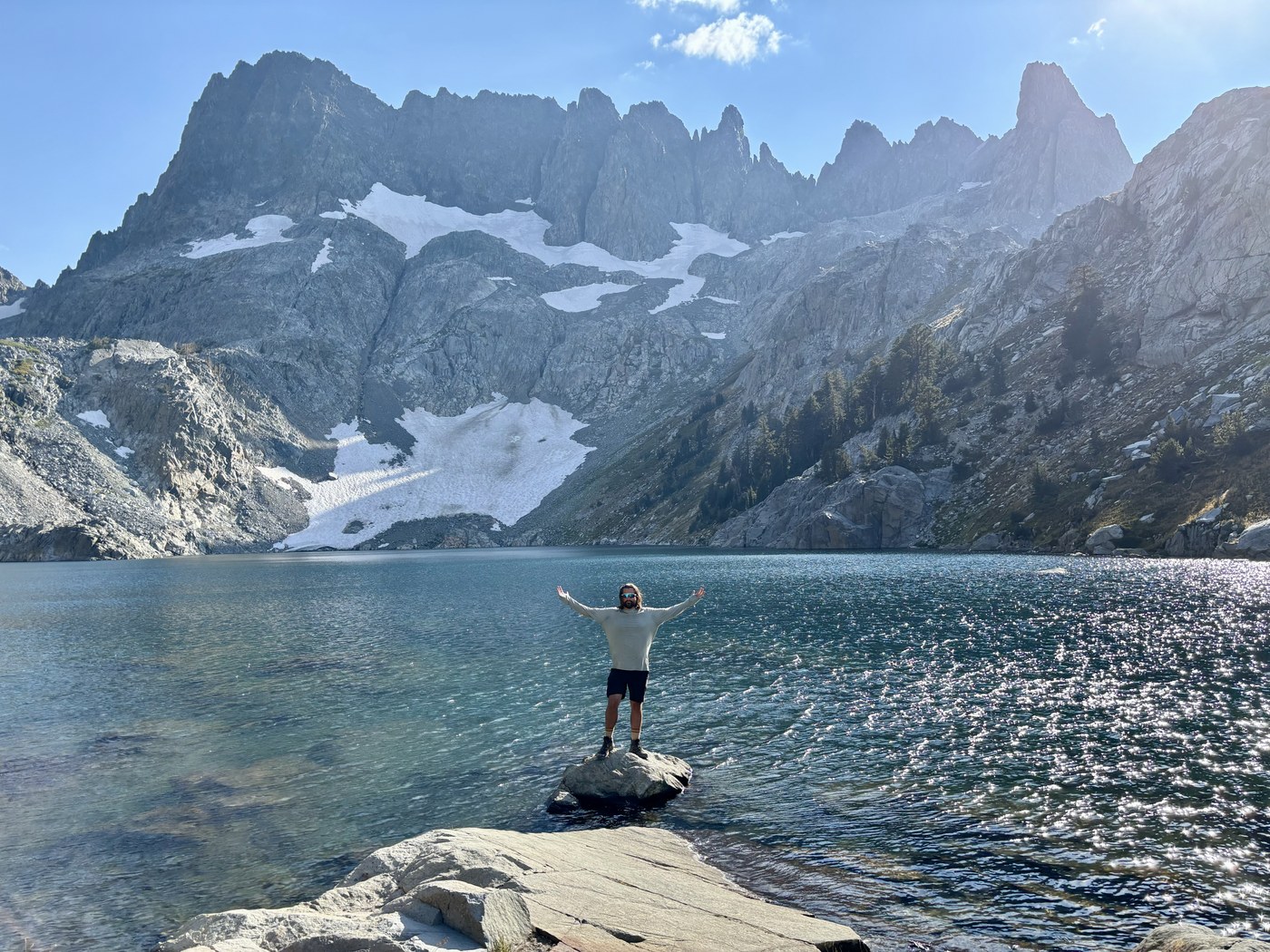 A hiker standing on a rock slab at the edge of a deep blue alpine lake with arms spread, a wall of jagged snow-streaked peaks rising directly behind the water