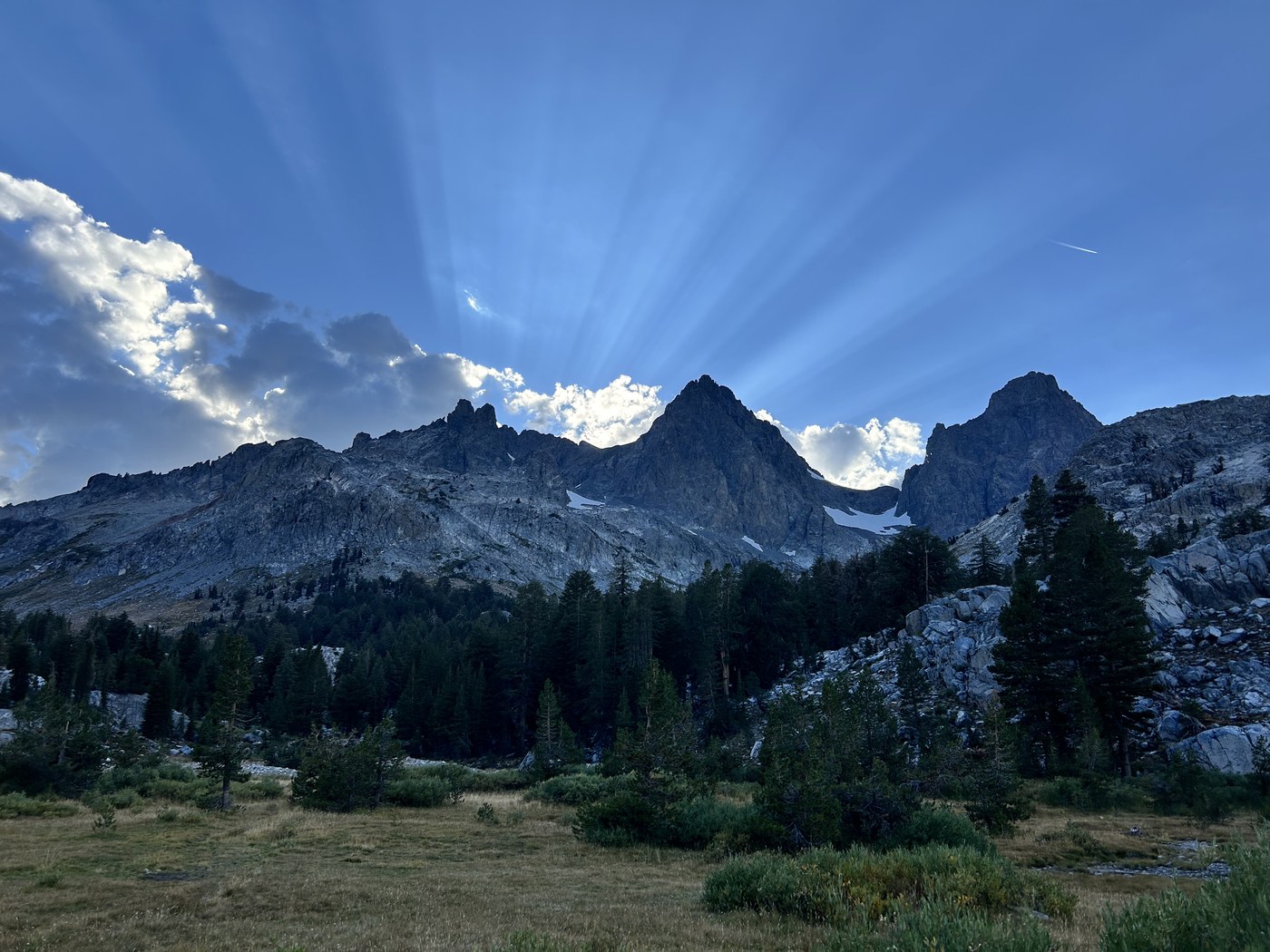 Long spokes of sunlight fanning out from behind a silhouetted ridge of granite spires above a dark meadow and treeline