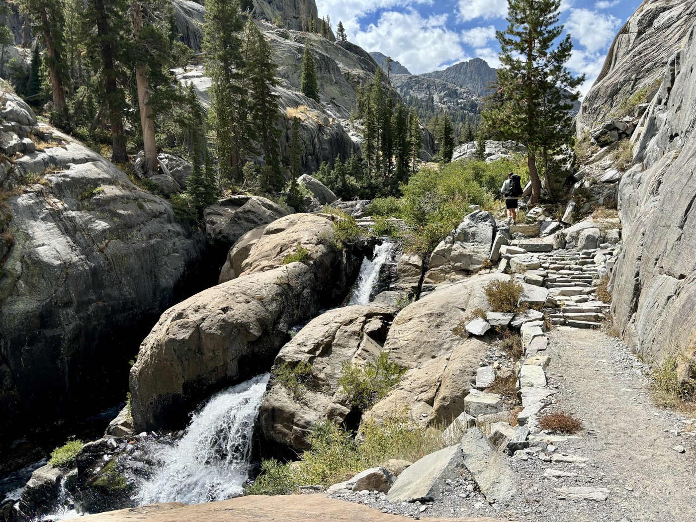 Water sliding and falling down bare pale granite beside a rocky trail climbing through scattered pines under a bright sky