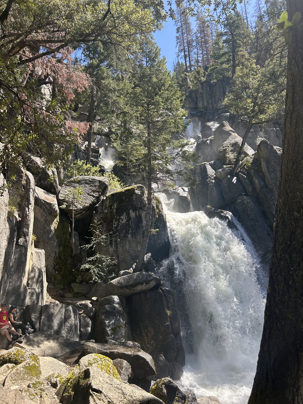 A powerful waterfall dropping through a granite gorge in full spring flow, with two small figures sitting on the rocks at the bottom left for scale