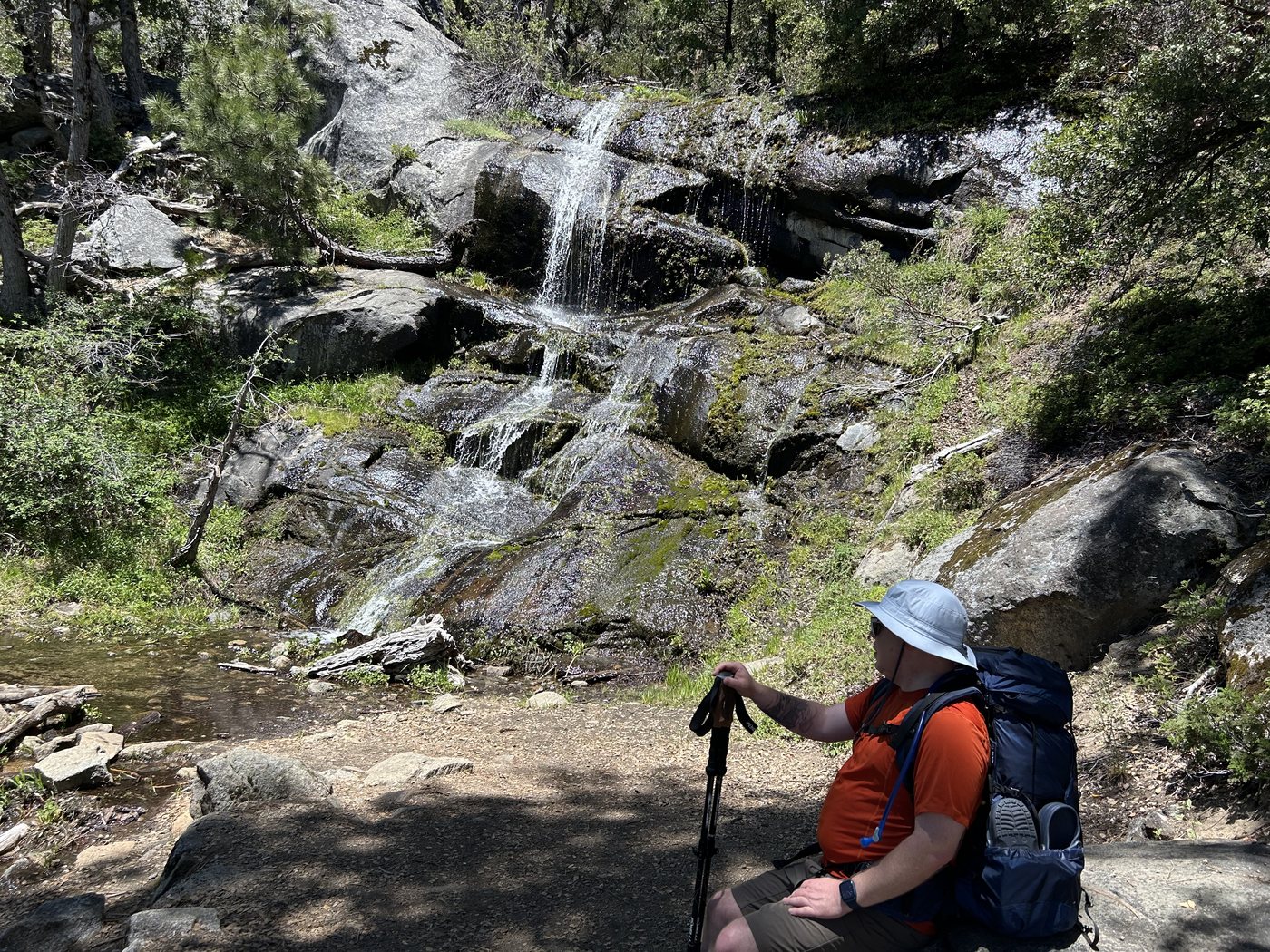 A hiker resting on a rock beside a thin waterfall spreading in several threads down a face of wet mossy granite