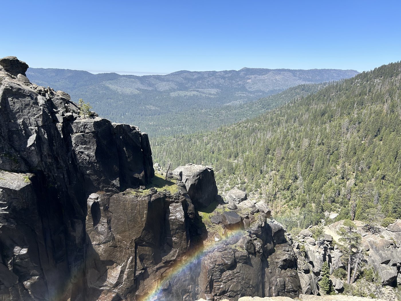 A rainbow standing in waterfall mist below dark cliffs, with a wide forested canyon and blue ridges beyond