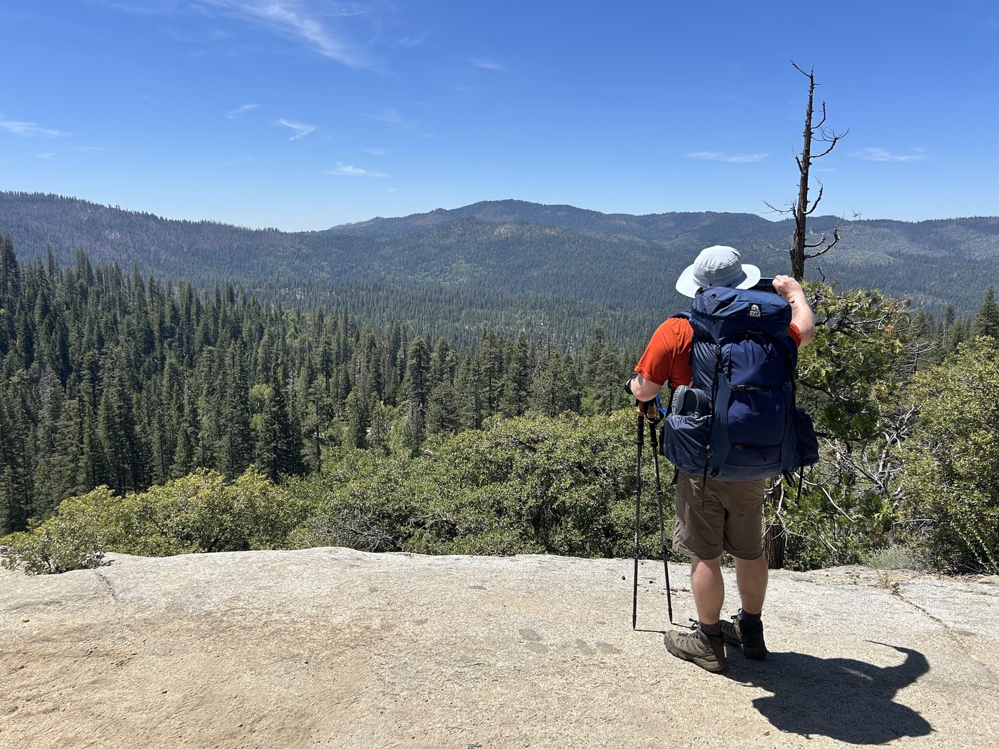 A backpacker with a navy pack and wide-brimmed sun hat standing on open granite slab, looking out over a forested basin toward distant ridges