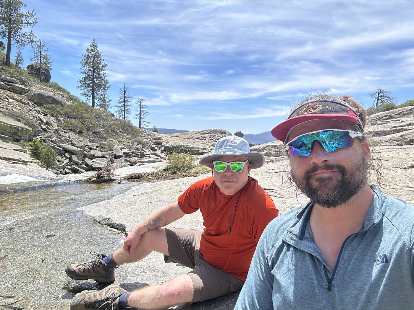 Two backpackers sitting side by side on a granite slab beside a rushing creek, both in sunglasses and squinting into bright sun