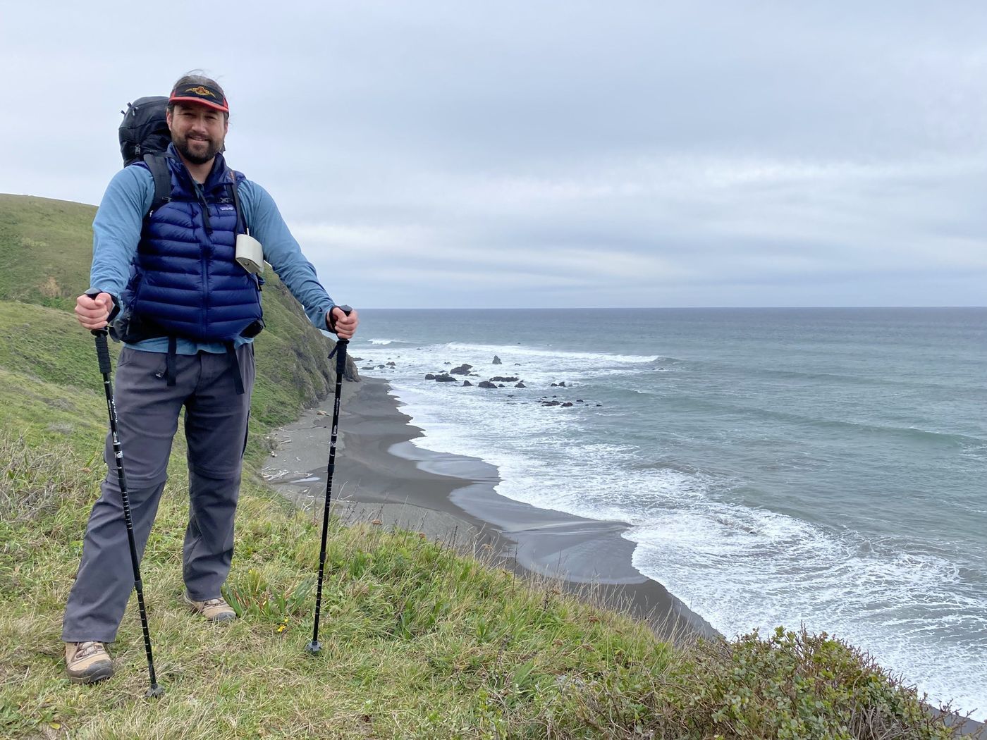 A backpacker standing on a high grassy bluff above the black sand beach and sea stacks of the Lost Coast