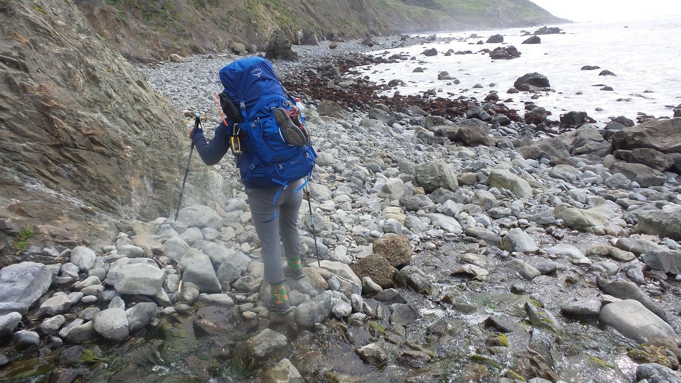 A backpacker picking a way across slick cobbles at the base of a cliff on the Lost Coast