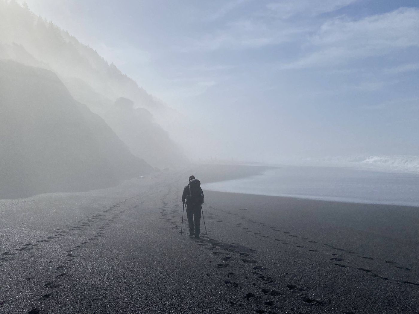 A lone backpacker walking into thick coastal fog on the black sand of the Lost Coast