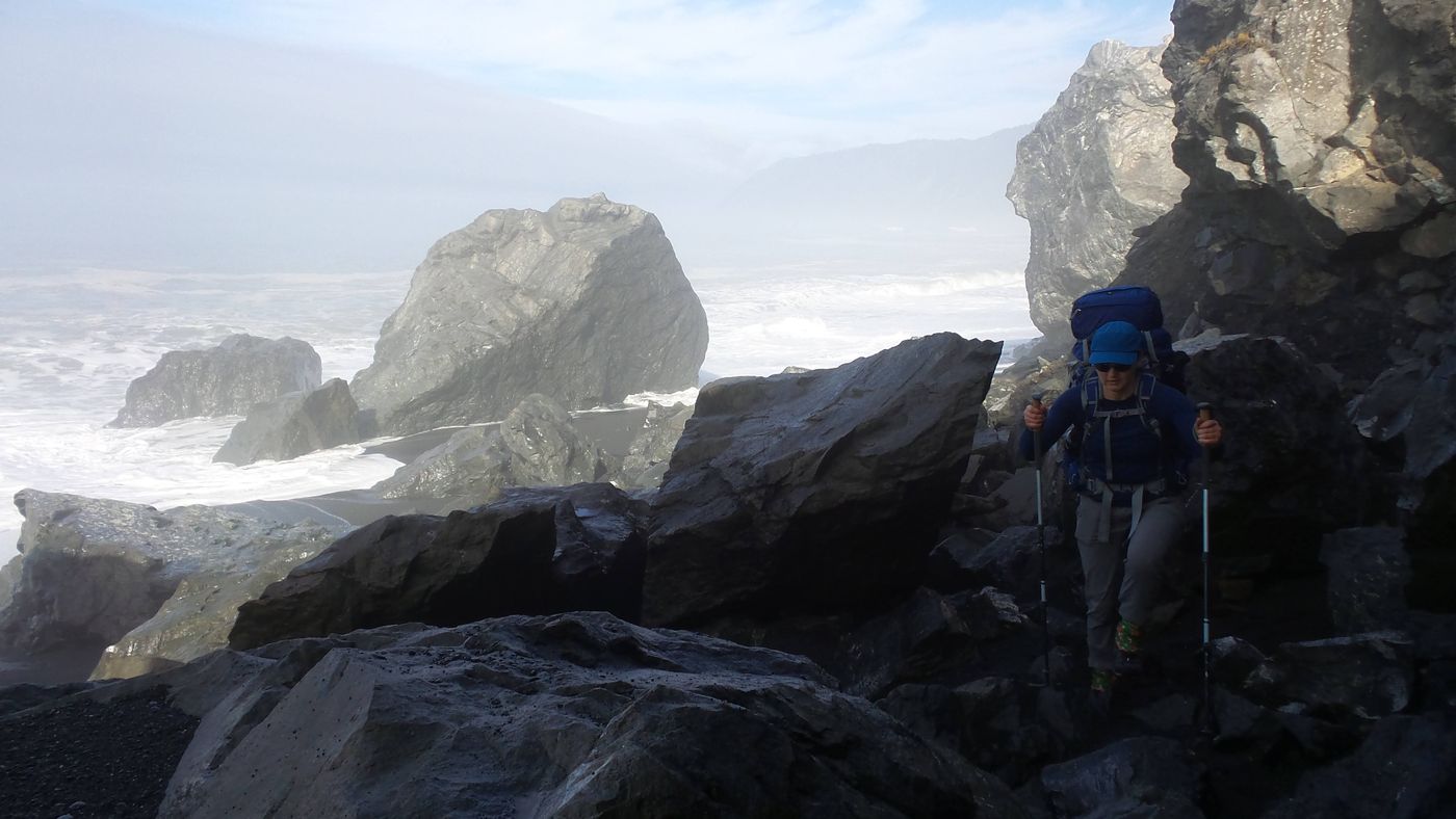 A backpacker scrambling among large sea stacks with surf breaking behind