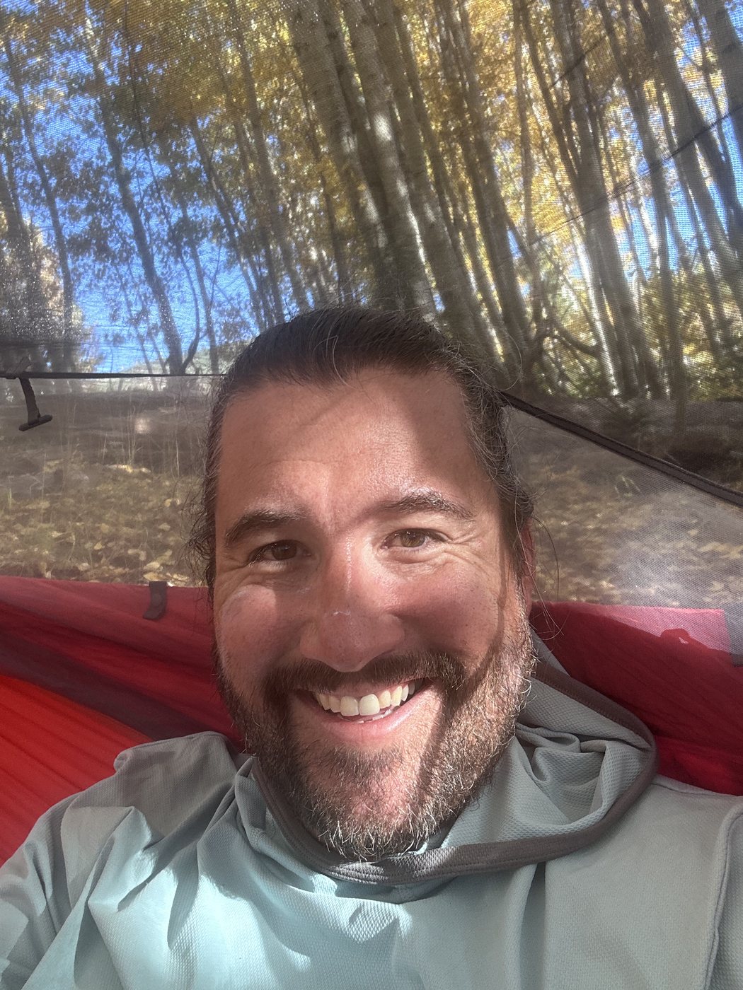 A smiling bearded man lying in a red hammock in a light hooded shirt, golden aspen trunks and leaves overhead