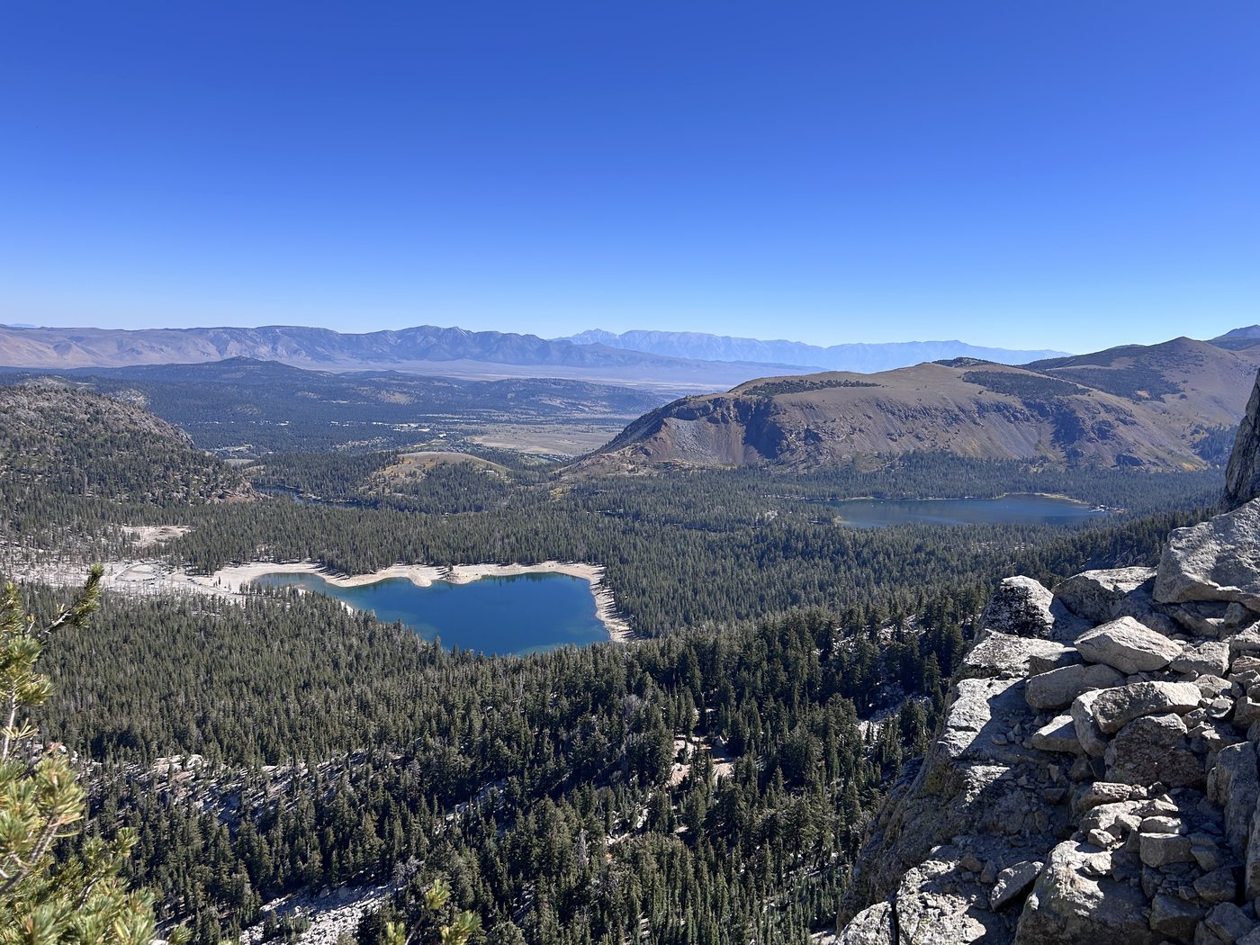 Looking down from a rocky crest over forest to a deep blue lake ringed with pale shoreline, more lakes and a broad valley stretching to distant desert ranges