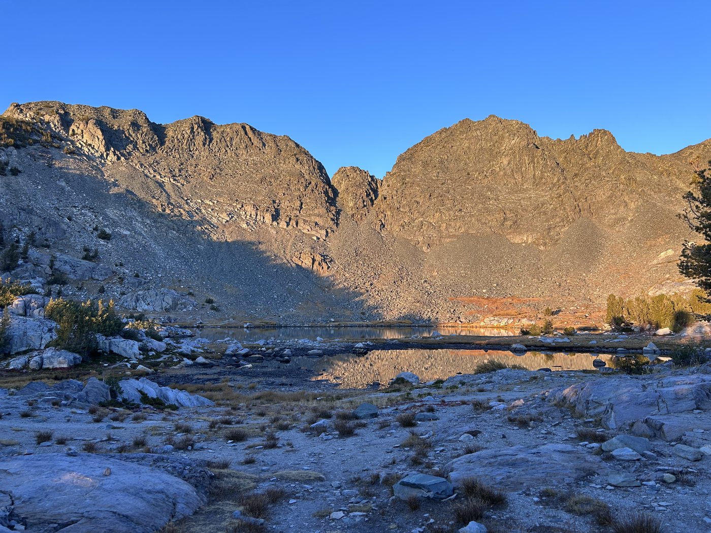 A granite cirque wall glowing orange in the last sun above a small dark lake and a foreground of pale boulders and low pines