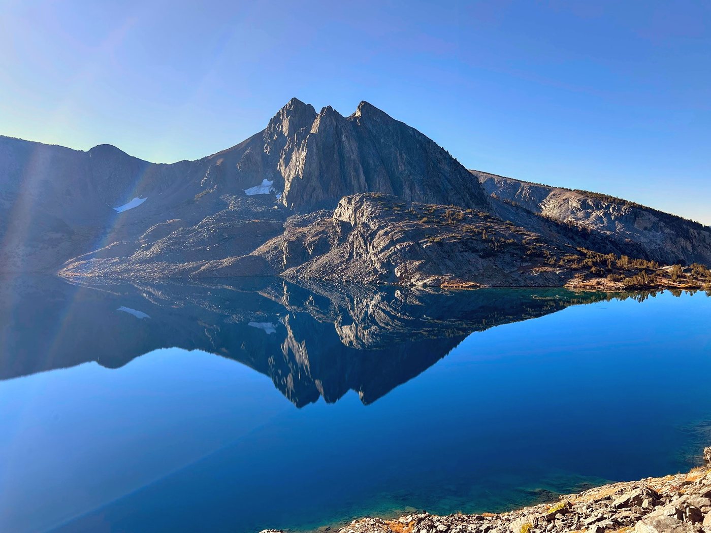 A twin-summited granite peak and the long ridge beside the peak reflected without a ripple in a deep blue alpine lake under a clear morning sky