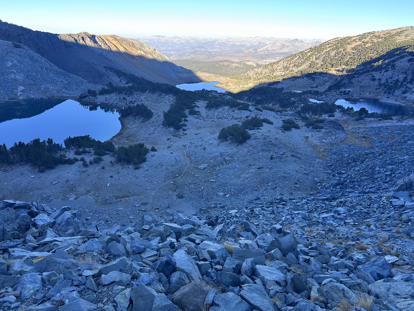 Looking back down a talus slope at small blue lakes in a rocky basin, the valley opening out to a wide hazy plain and distant ranges