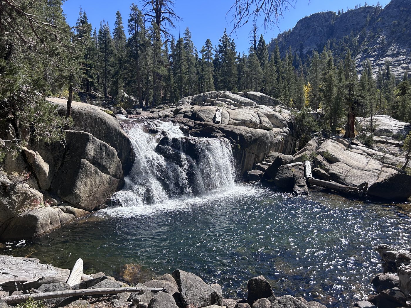 A creek pouring over a short granite ledge into a round clear pool ringed with boulders, pines on the far bank under a blue sky