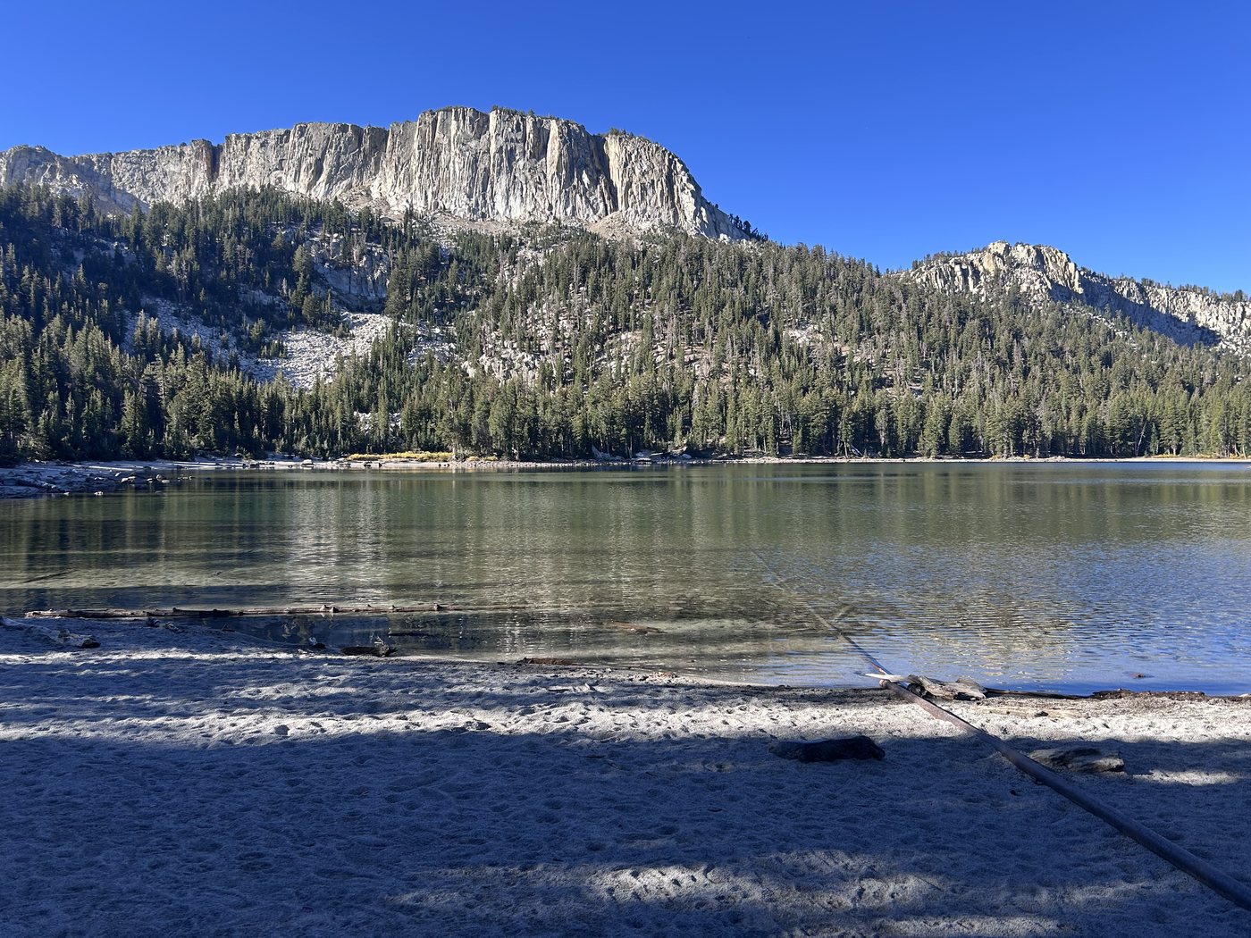 A calm lake with a pale sandy shore in morning shadow, a long gray cliff band rising above forested slopes on the far side under a deep blue sky