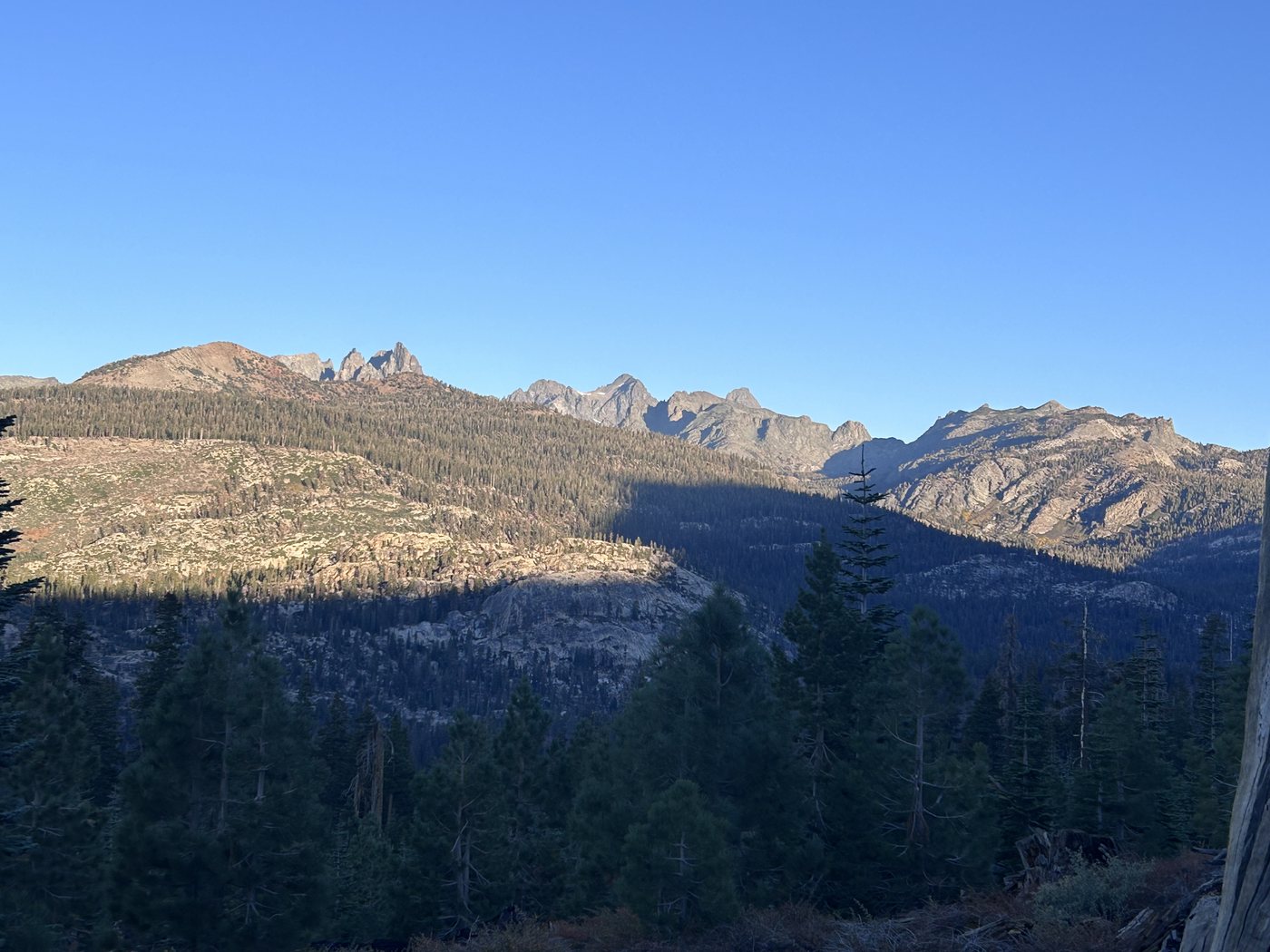 A jagged range of dark spires lit by early sun on the horizon, above a long forested valley still in shadow