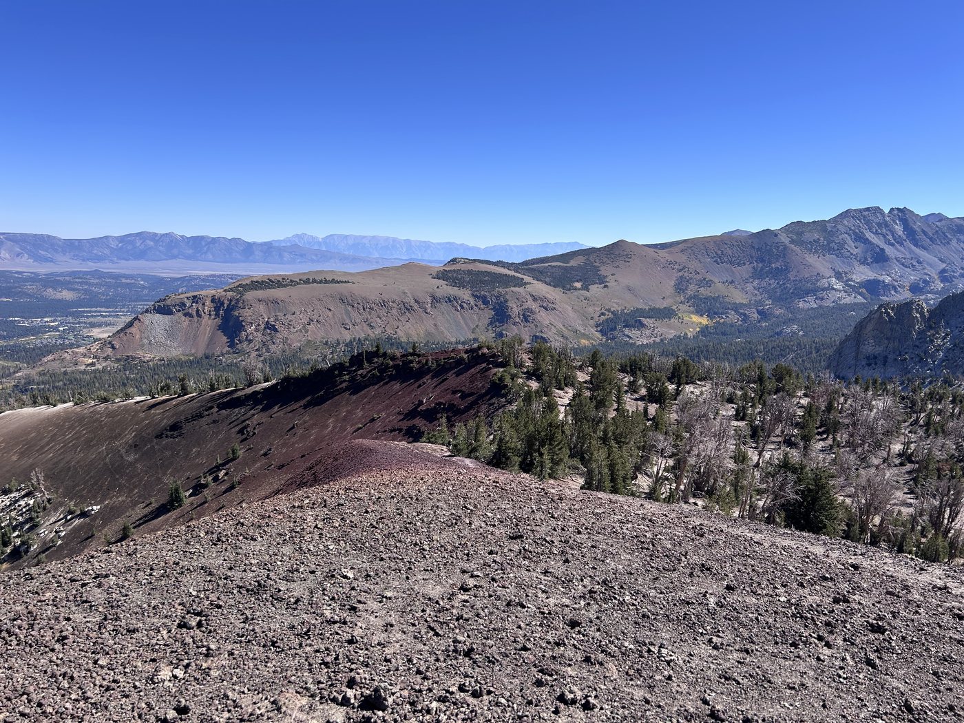 A broad slope of dark brown cinder in the foreground, a band of red volcanic ground beyond, and a range of jagged gray peaks across a forested basin