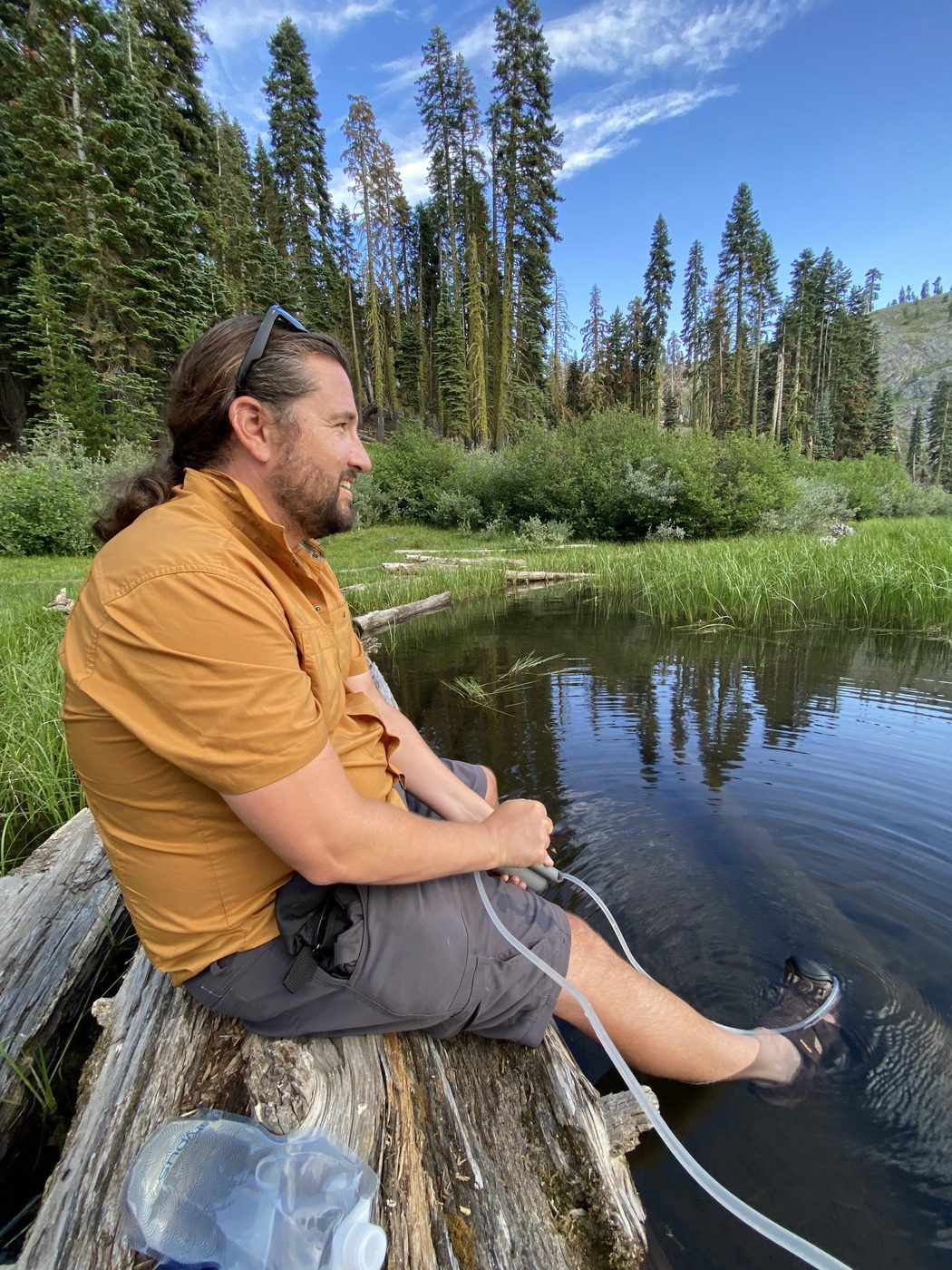 A man in an orange shirt sitting on a log at the water's edge, feet in the lake, filtering water and smiling
