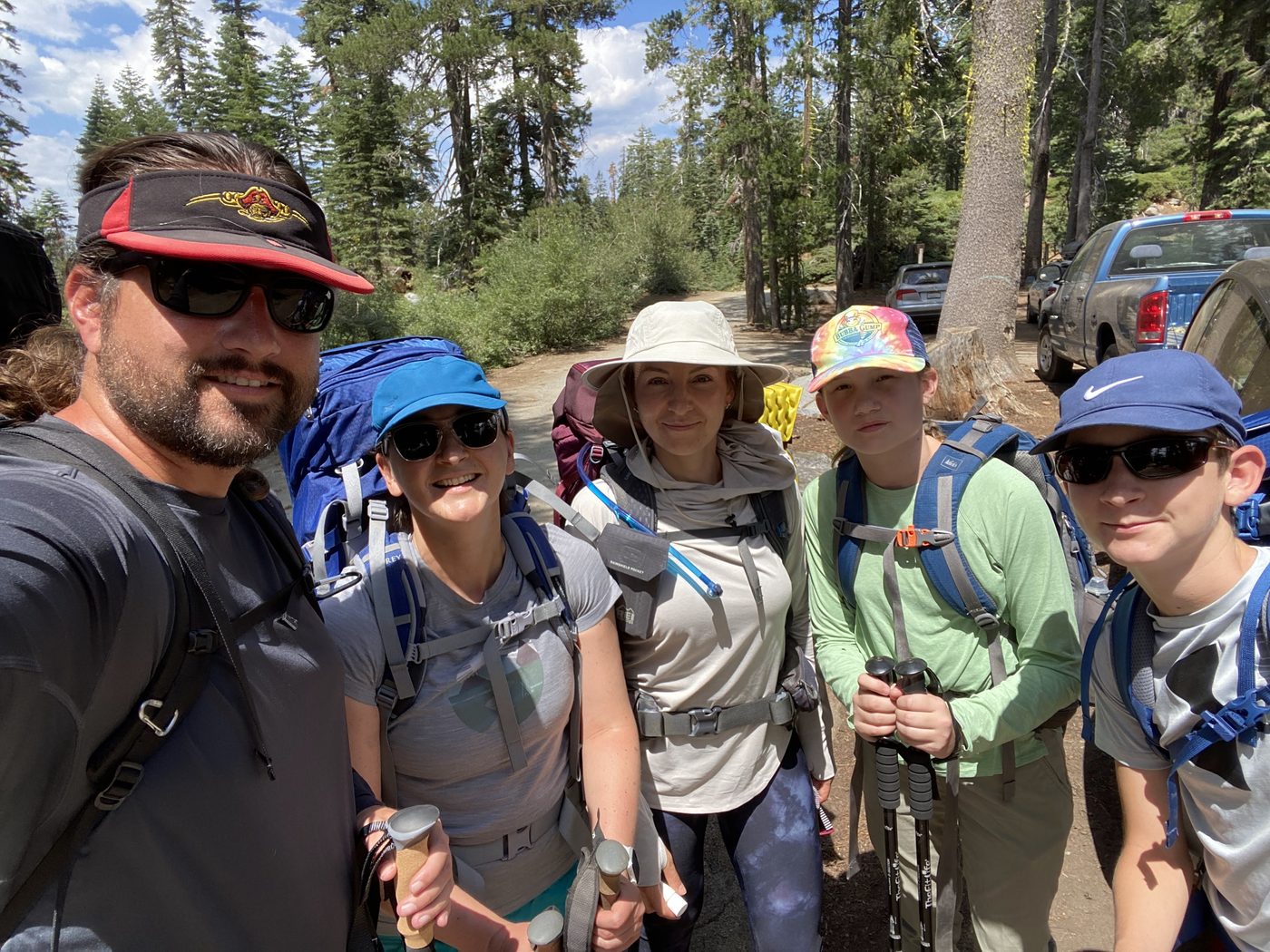A group selfie of five people in sun hats and full backpacks at a forested trailhead