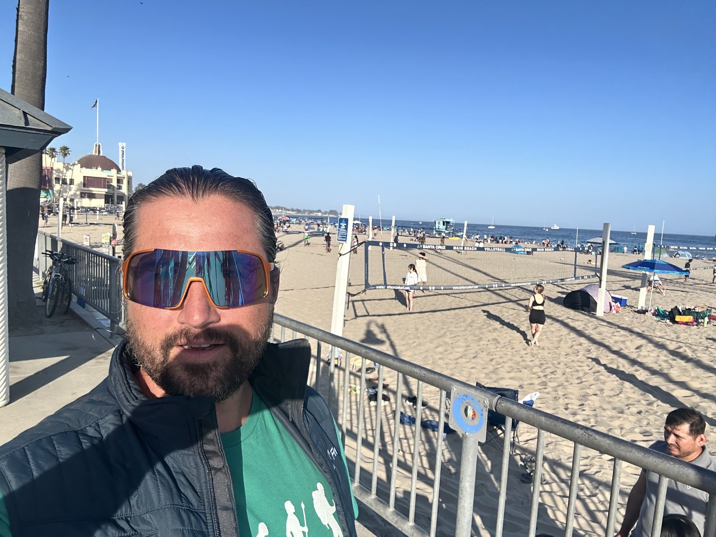 A close selfie against a beach railing with sand, volleyball nets, and the Pacific Ocean visible behind, evening light and a line of colorful beach houses in the distance