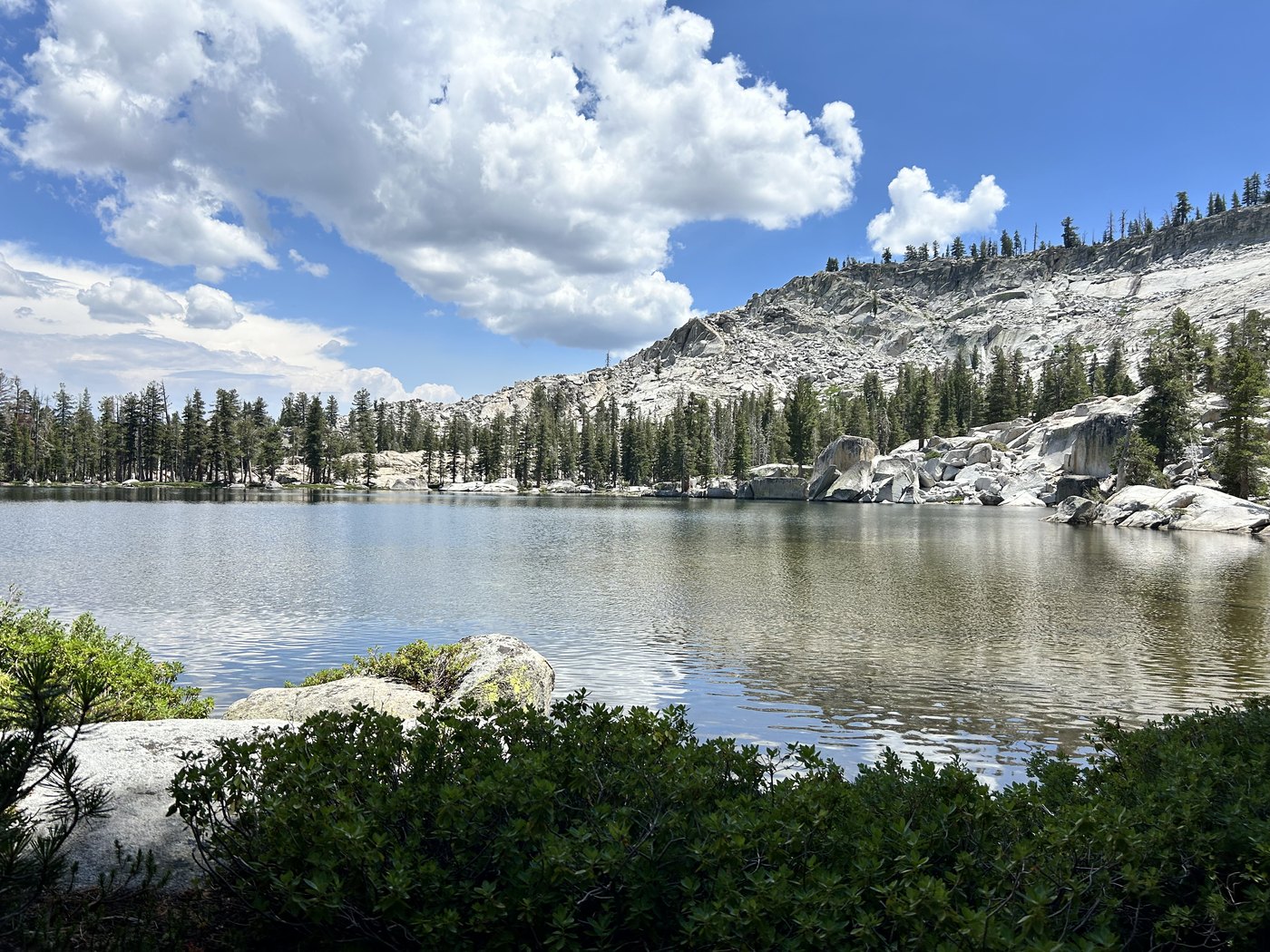 A calm alpine lake reflecting a pale granite dome and a partly cloudy sky, low green shrubs growing along the near shore