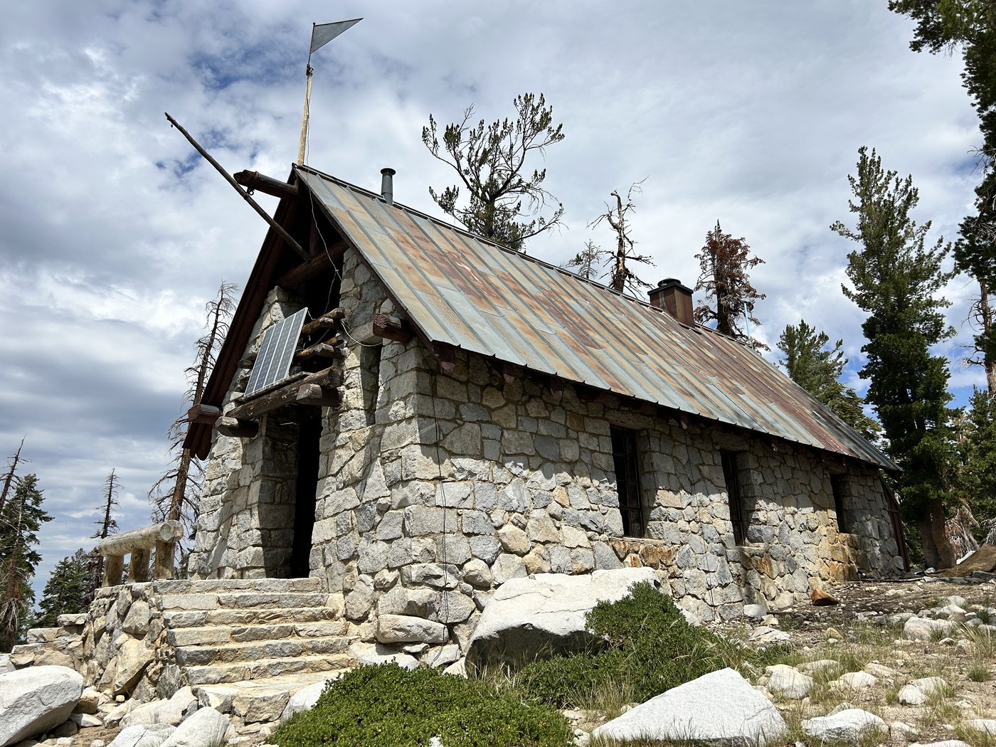 A low stone building with a rust-streaked metal roof and a small solar panel above the door, reached by a stone stairway, standing alone among pines under a cloudy sky