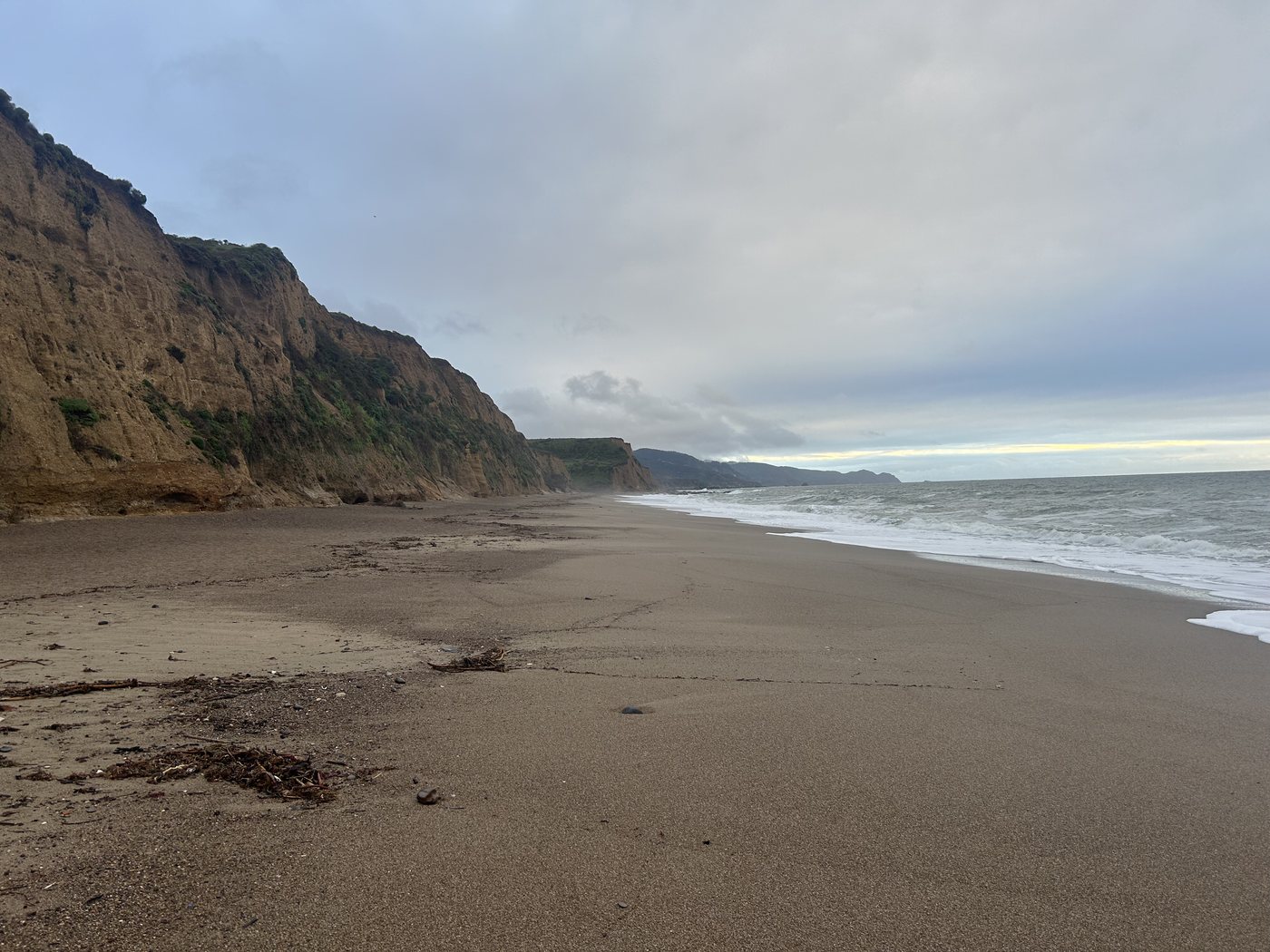A wide empty beach at low tide running south beneath tall eroding cliffs under an overcast sky