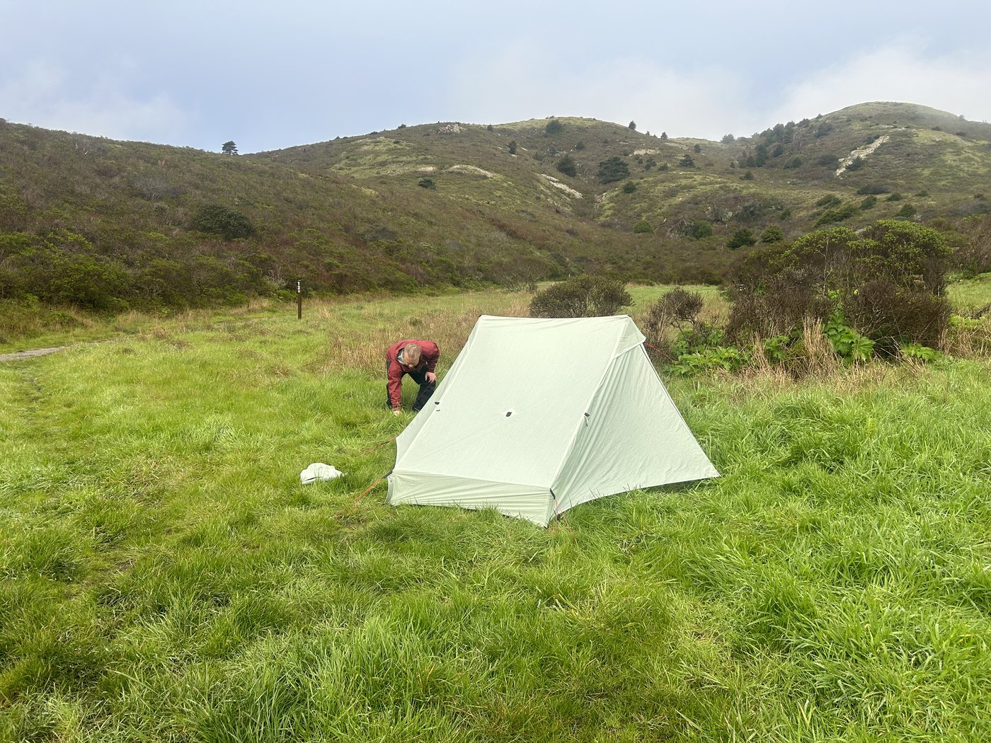 A tan A-frame trekking-pole tent pitched in bright green grass in an open bowl below rounded coastal hills, a hiker in a red jacket staking it out