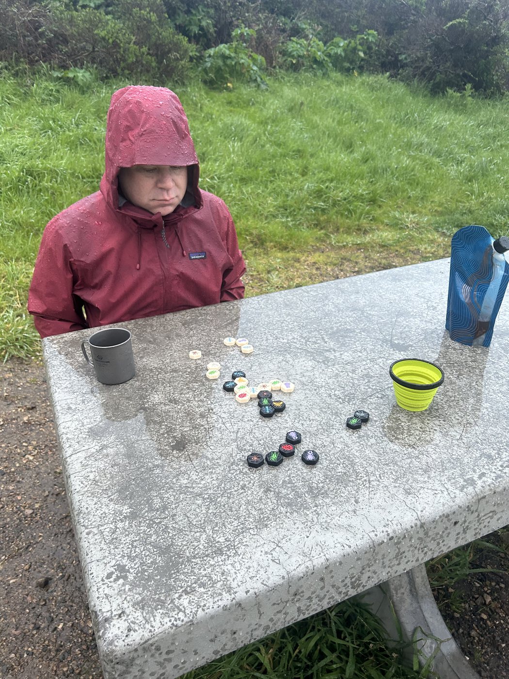 A hiker in a soaked red rain shell with the hood up, sitting at a wet concrete picnic table studying a game of Hive laid out in front of him