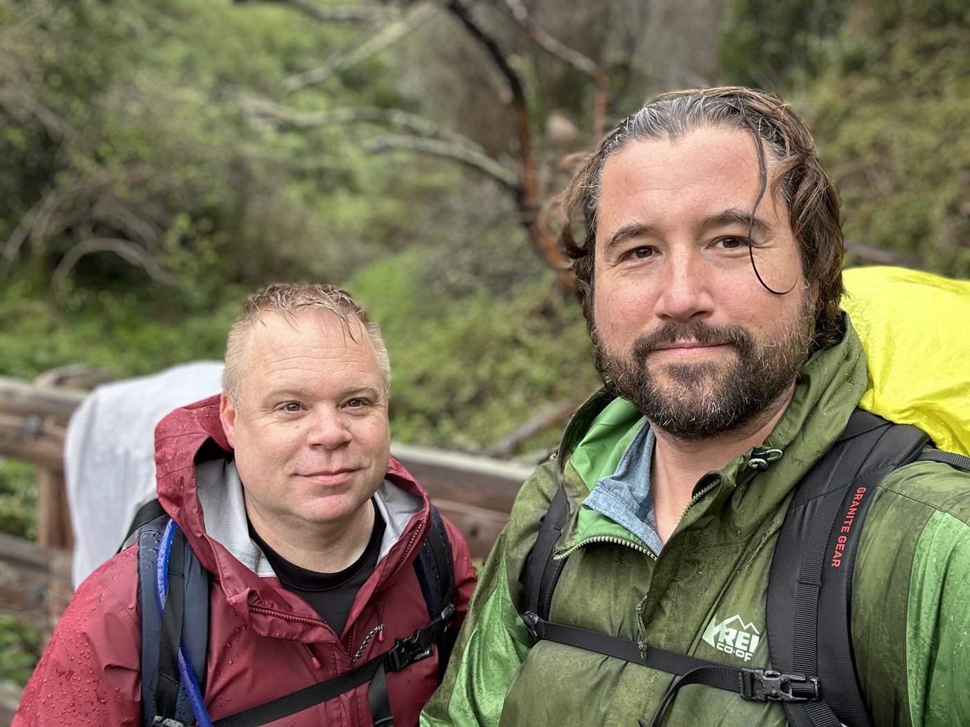 Two backpackers with soaked hair standing close together on a wet trail, one in a red rain shell and one in a green rain shell with a yellow pack cover