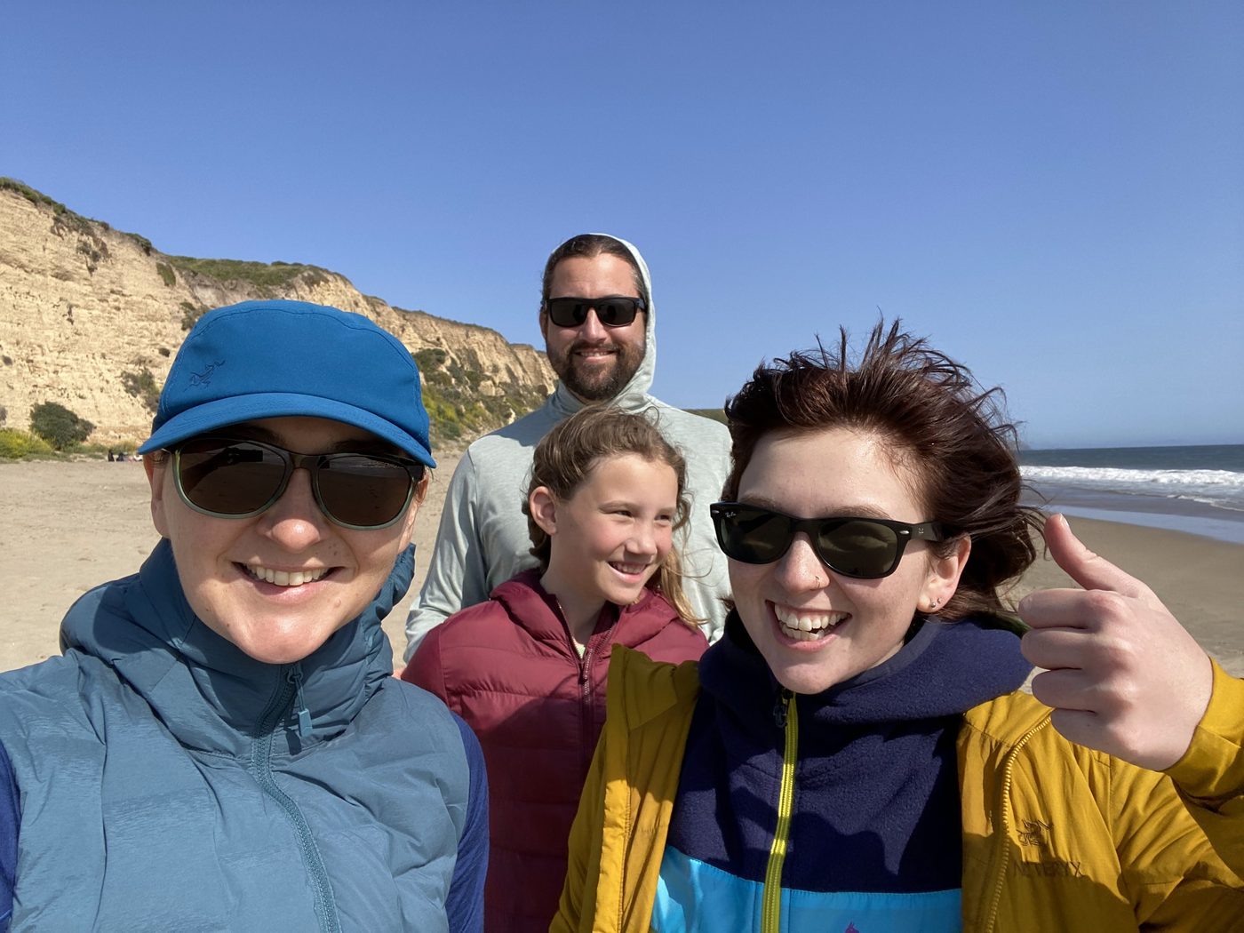 A bright group selfie of four people on the beach, cliffs and surf behind them, everyone smiling