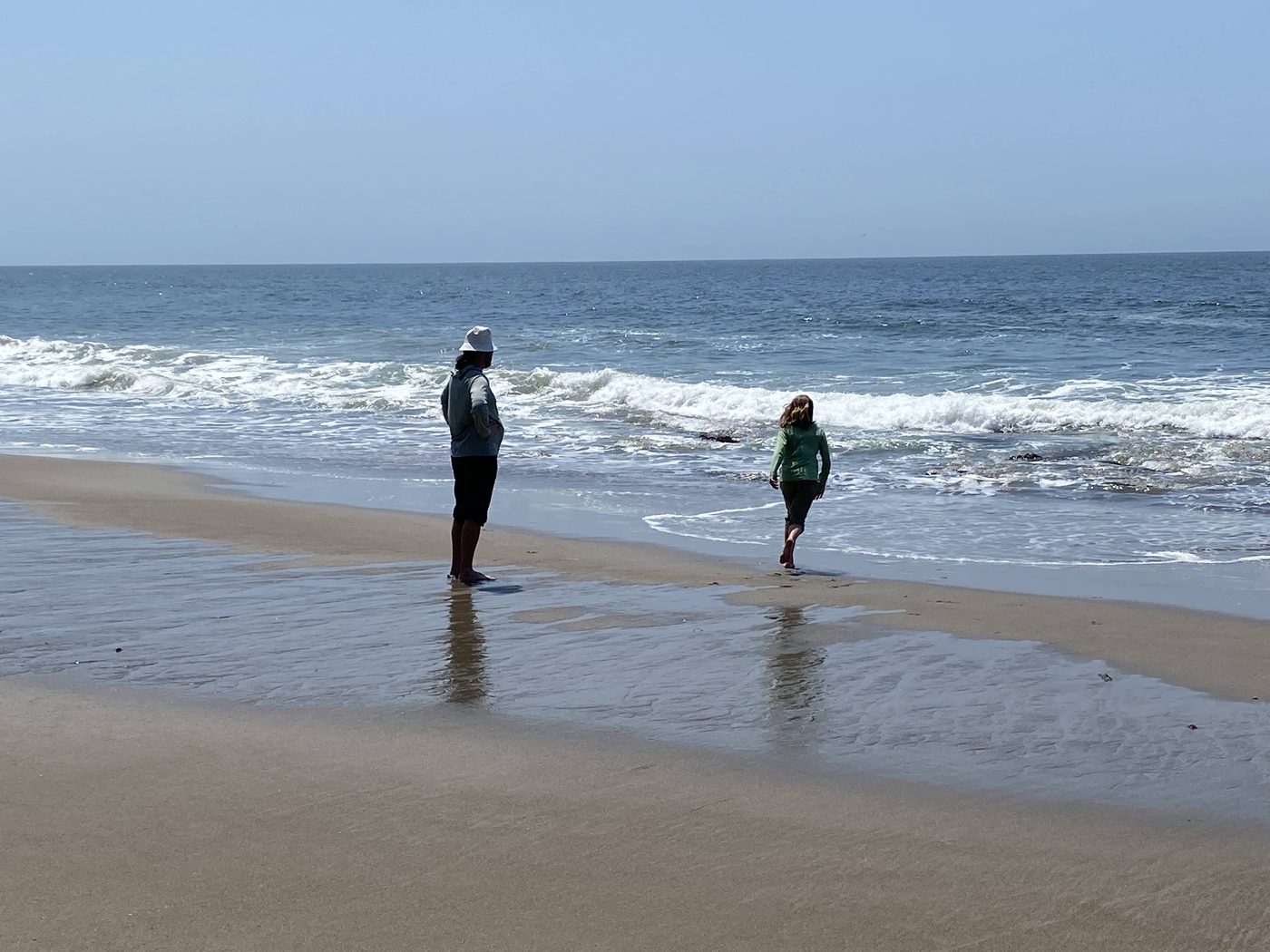 A woman and a girl standing at the edge of the surf on a wide empty beach
