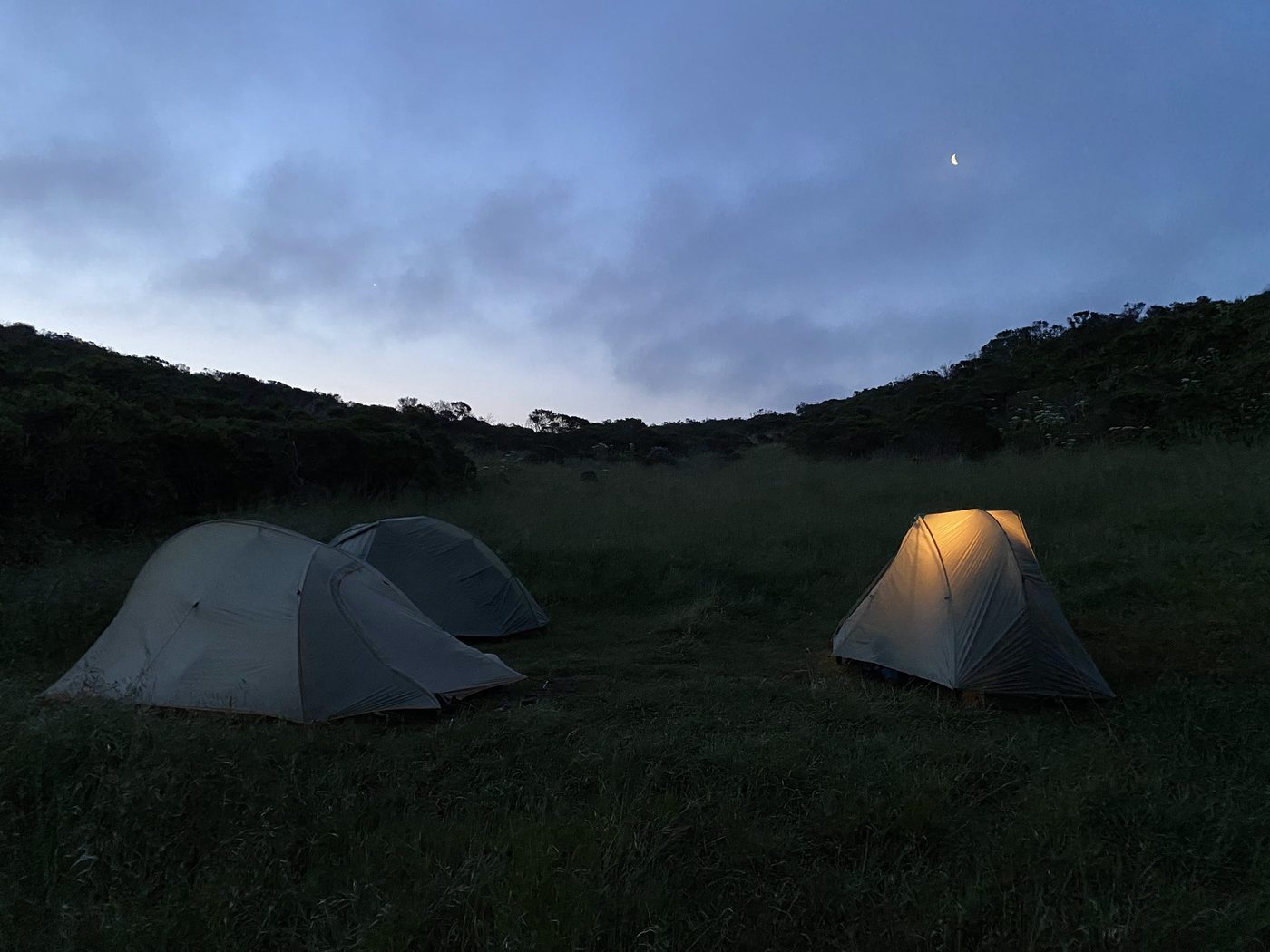 Two backpacking tents in a grassy meadow at dusk, a crescent moon above, one tent glowing from within