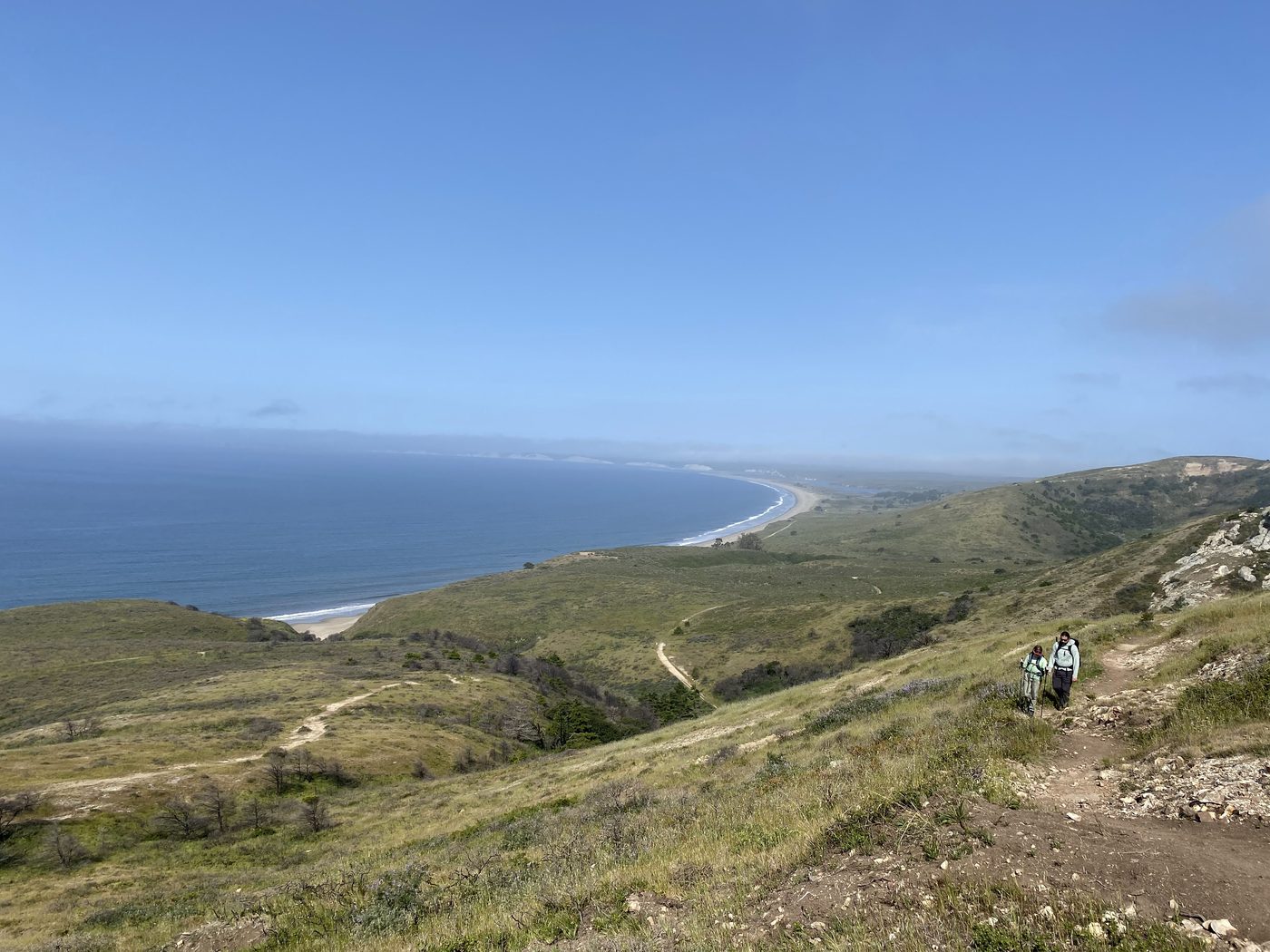 Two small hikers on a trail climbing a green ridge high above a long curving beach and bay