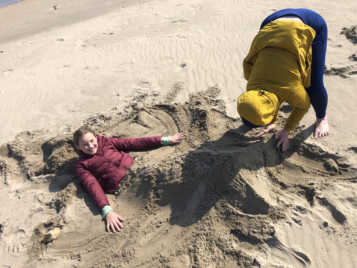 A girl in a maroon jacket lying in the sand, smiling, while an adult sculpts a sand creature around her