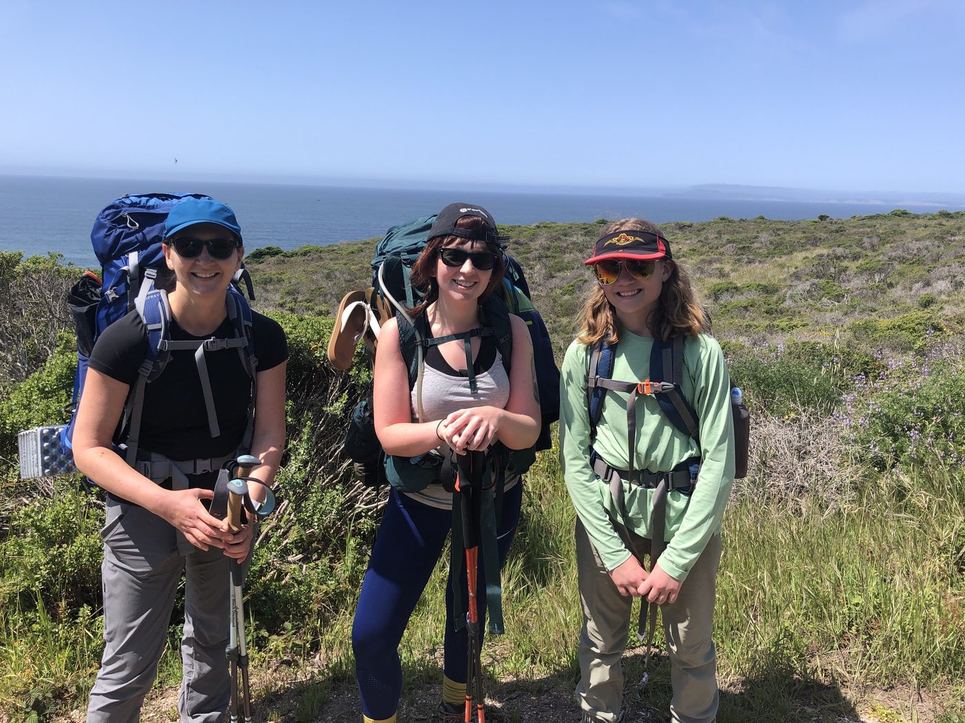 Three female backpackers with full packs and trekking poles standing on a coastal bluff trail, the Pacific behind them