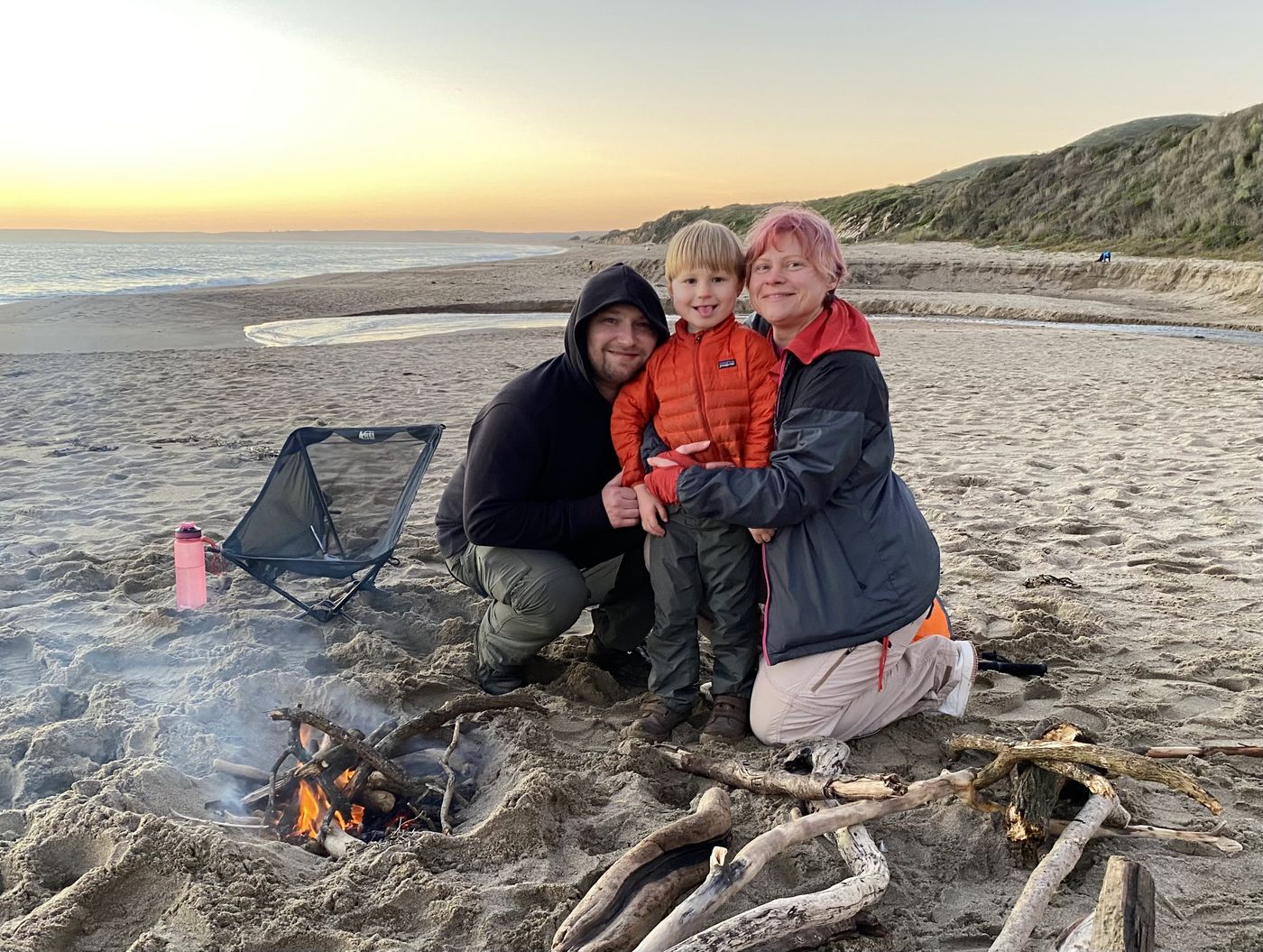 A family and a small child gathered around a small driftwood fire on the beach at sunset