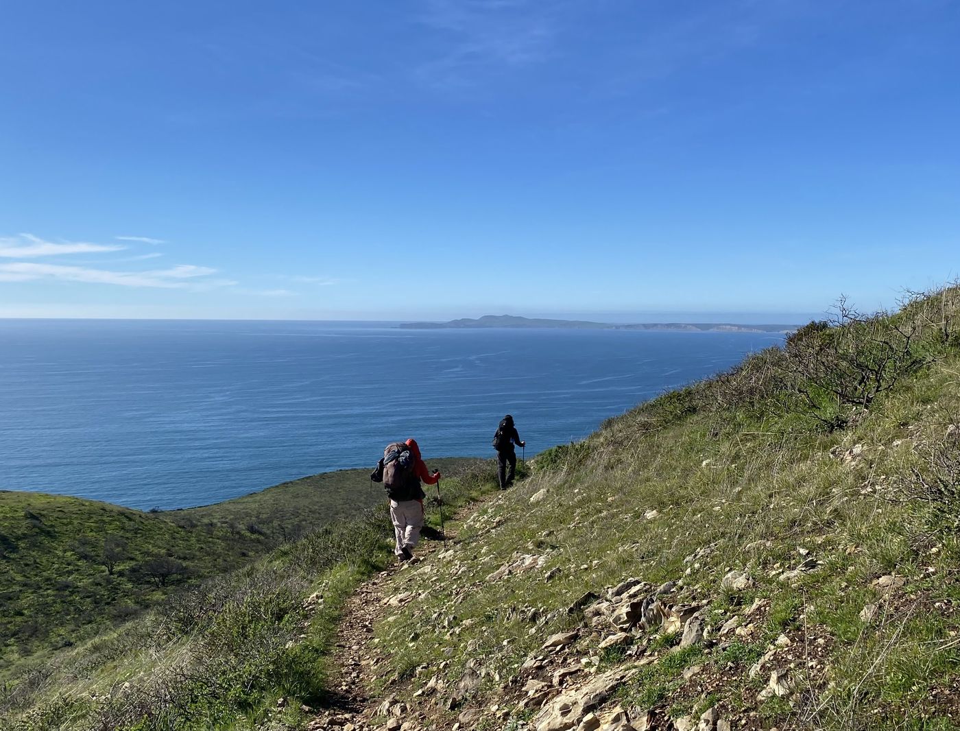 Two backpackers descending a grassy bluff trail toward the wide blue water of Drakes Bay