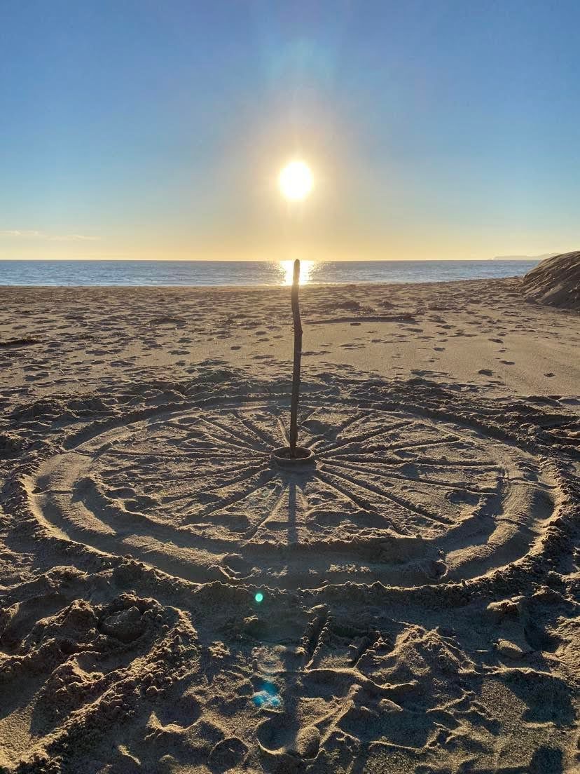 A large sun-wheel drawn in the beach sand with a driftwood stick at the center, the setting sun behind it
