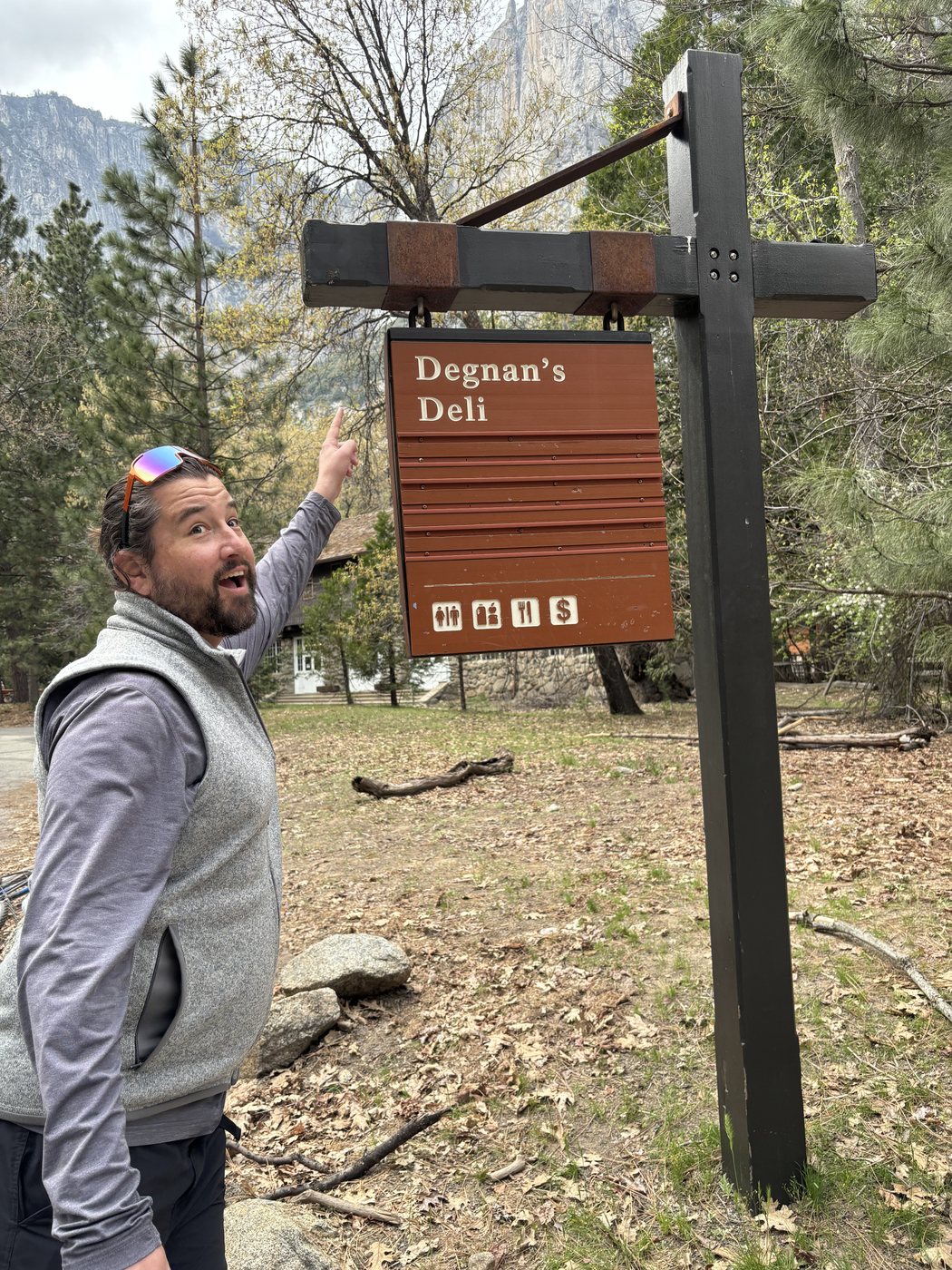 A hiker in a grey fleece vest pointing up at a brown national park sign reading Degnan's Deli, with an open-mouthed delighted expression